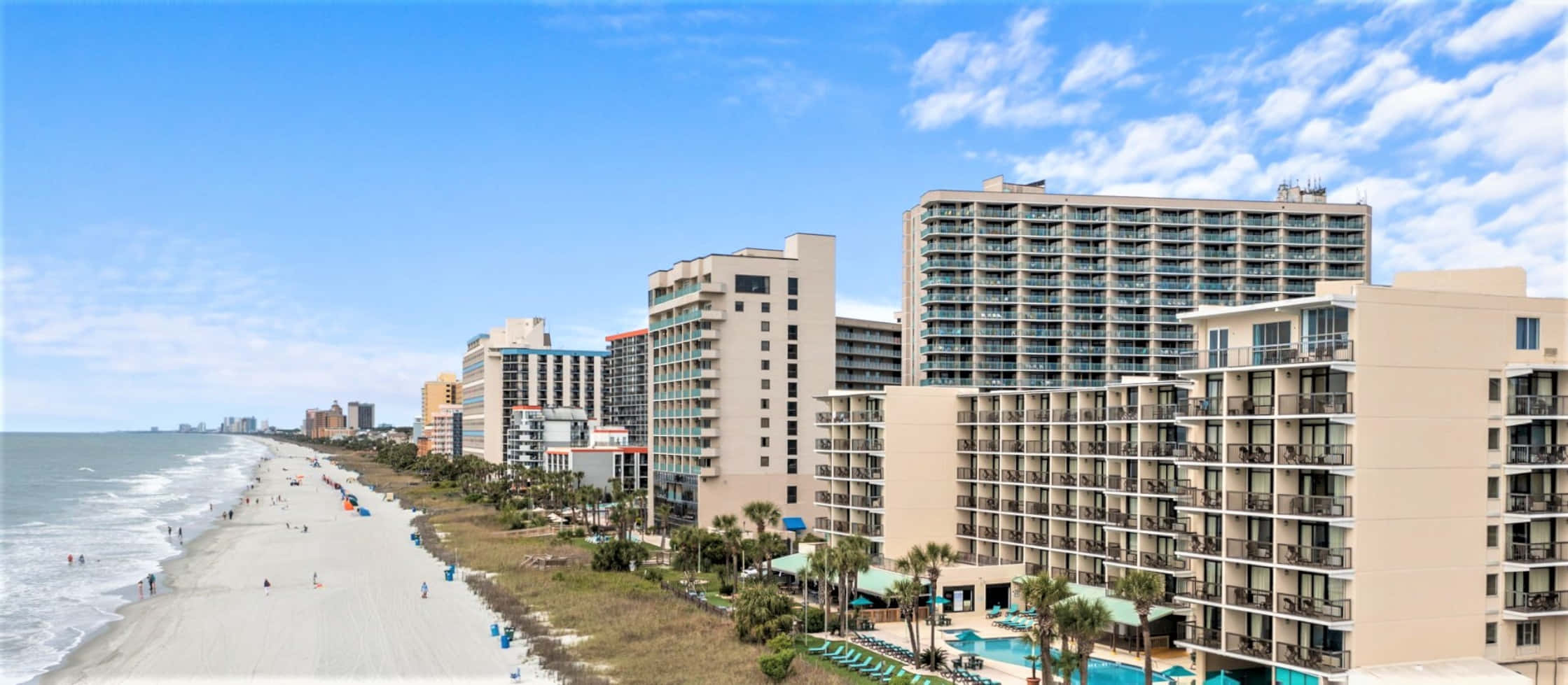 Myrtle Beach 桌布 壁紙 — 陽光明媚的海濱，高聳的飯店和棕櫚樹林立，天空湛藍，白雲點綴。