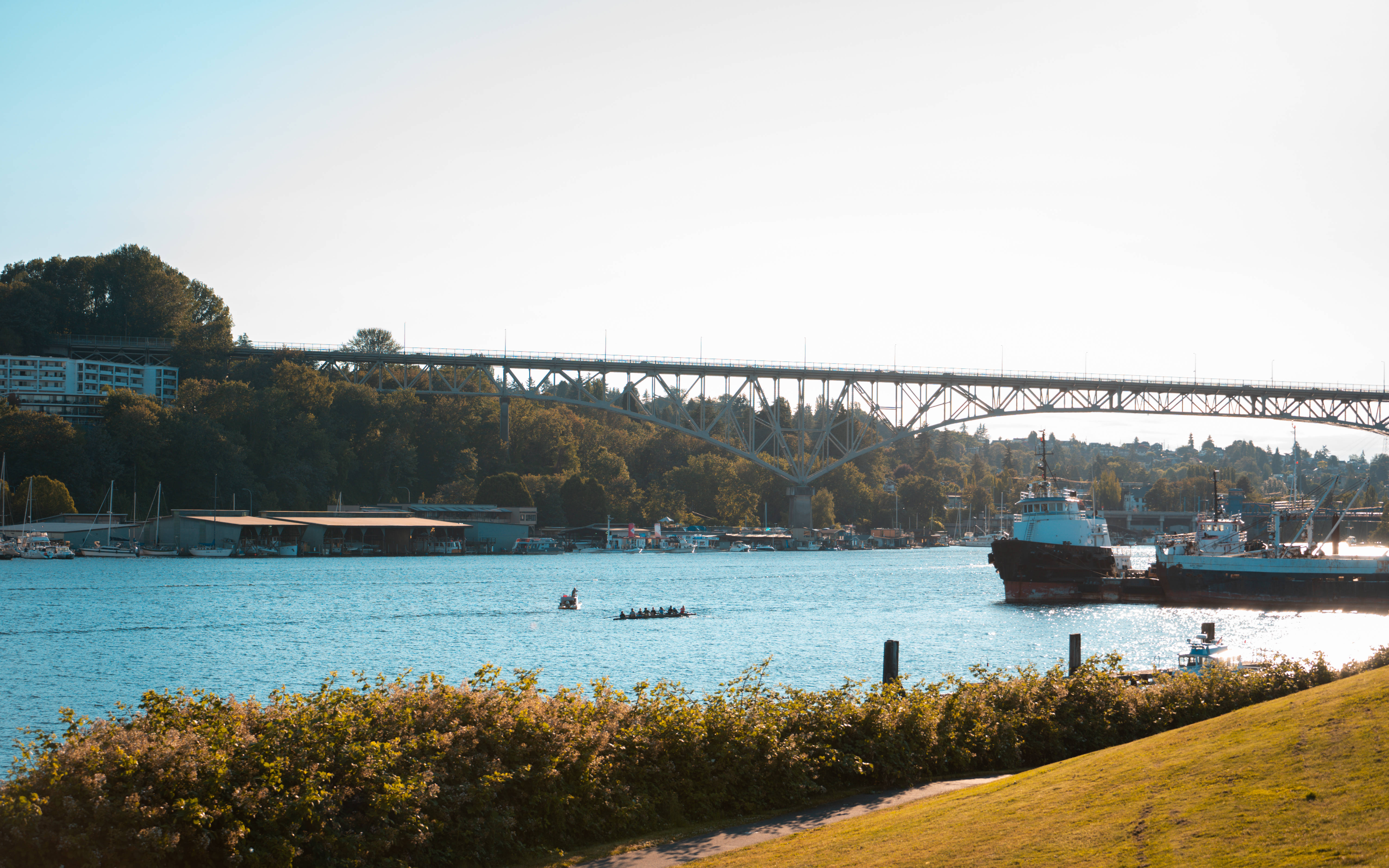 seattle 4K 桌布 壁紙 — 陽光明媚的海濱場景，一座橋橫跨平靜的藍色水道，船隻和划槳者滑過茂密的綠植和遠處的建築物。