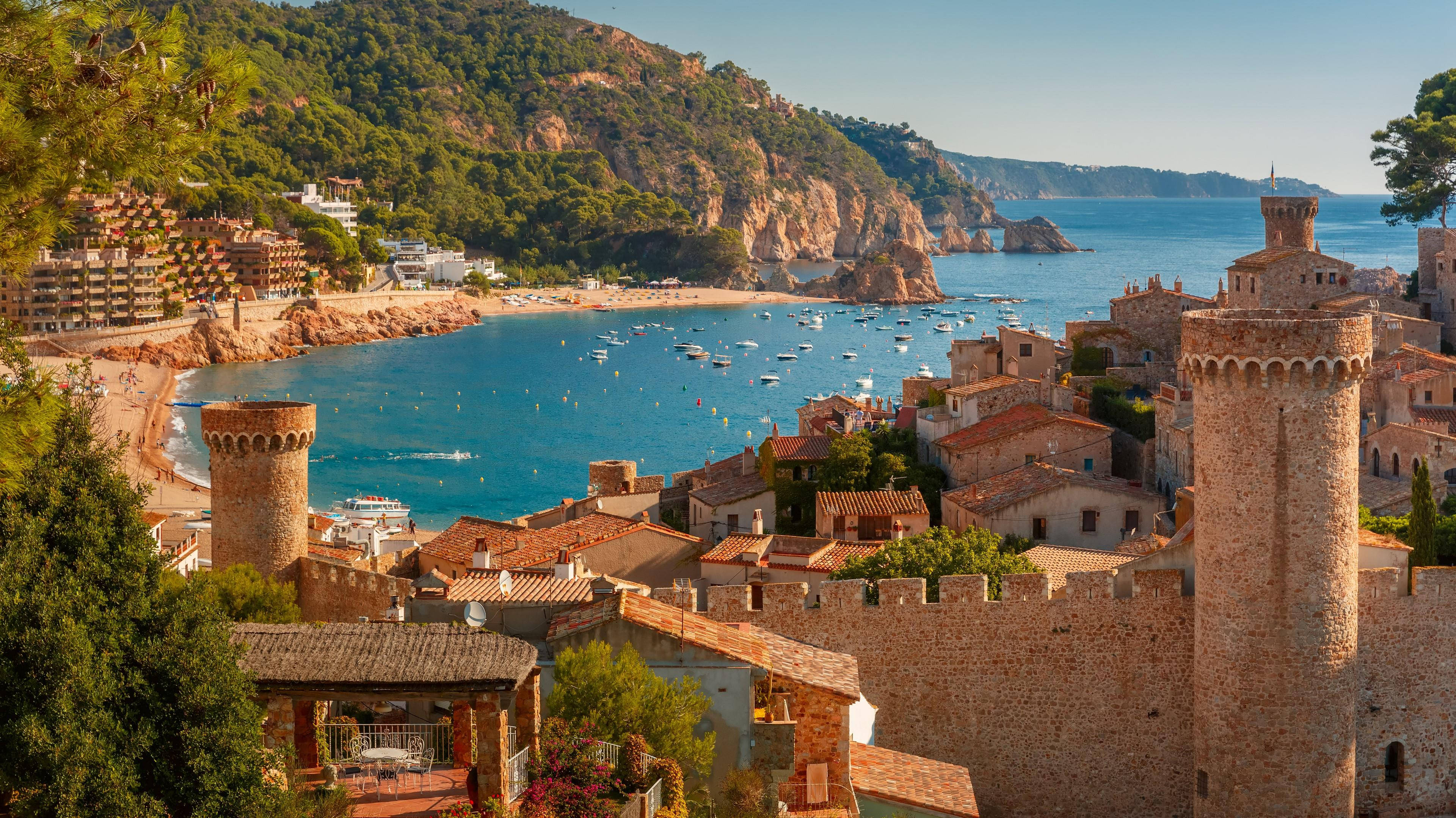 Spain 桌布 壁紙 — 陽光明媚的海濱小鎮，擁有陶土屋頂和石塔，毗鄰點綴著船隻的碧綠海水。