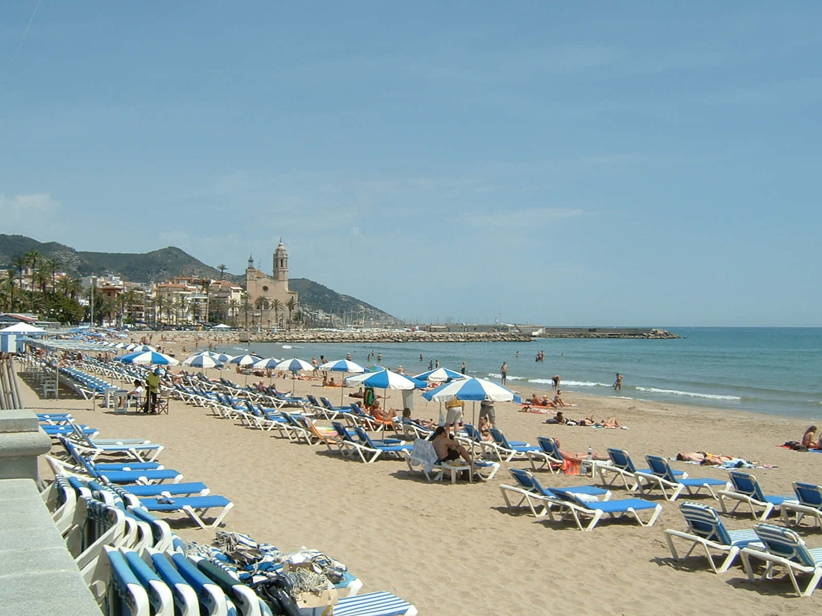 Spanish Beach 桌布 壁紙 — 陽光明媚的海灘，佈滿藍色躺椅和條紋遮陽傘，天空晴朗，遠處有教堂和山丘。