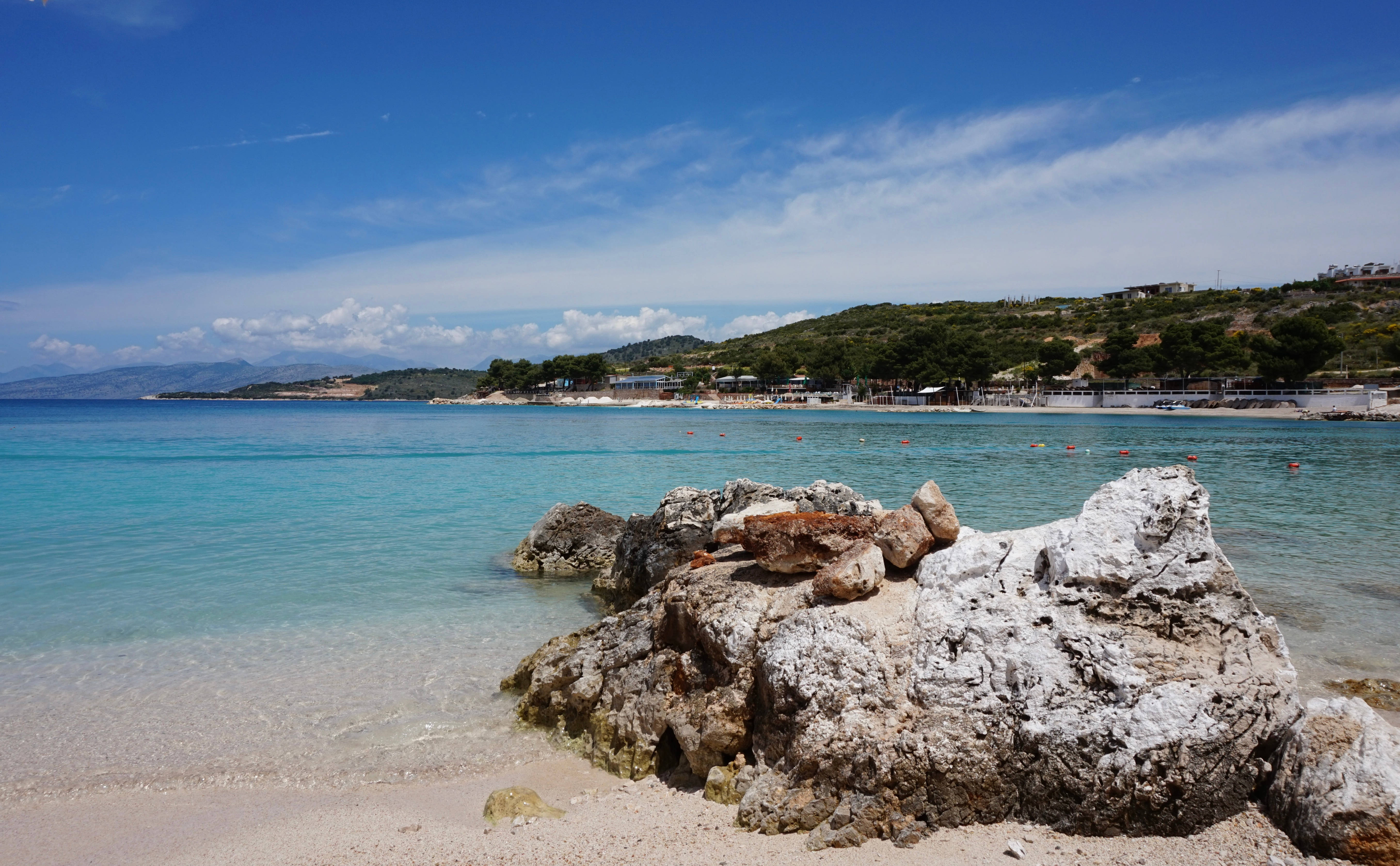 Albania 桌布 壁紙 — 陽光明媚的海灘，前景有碧綠的海水和大型紋理岩石，上方是明亮的藍天。