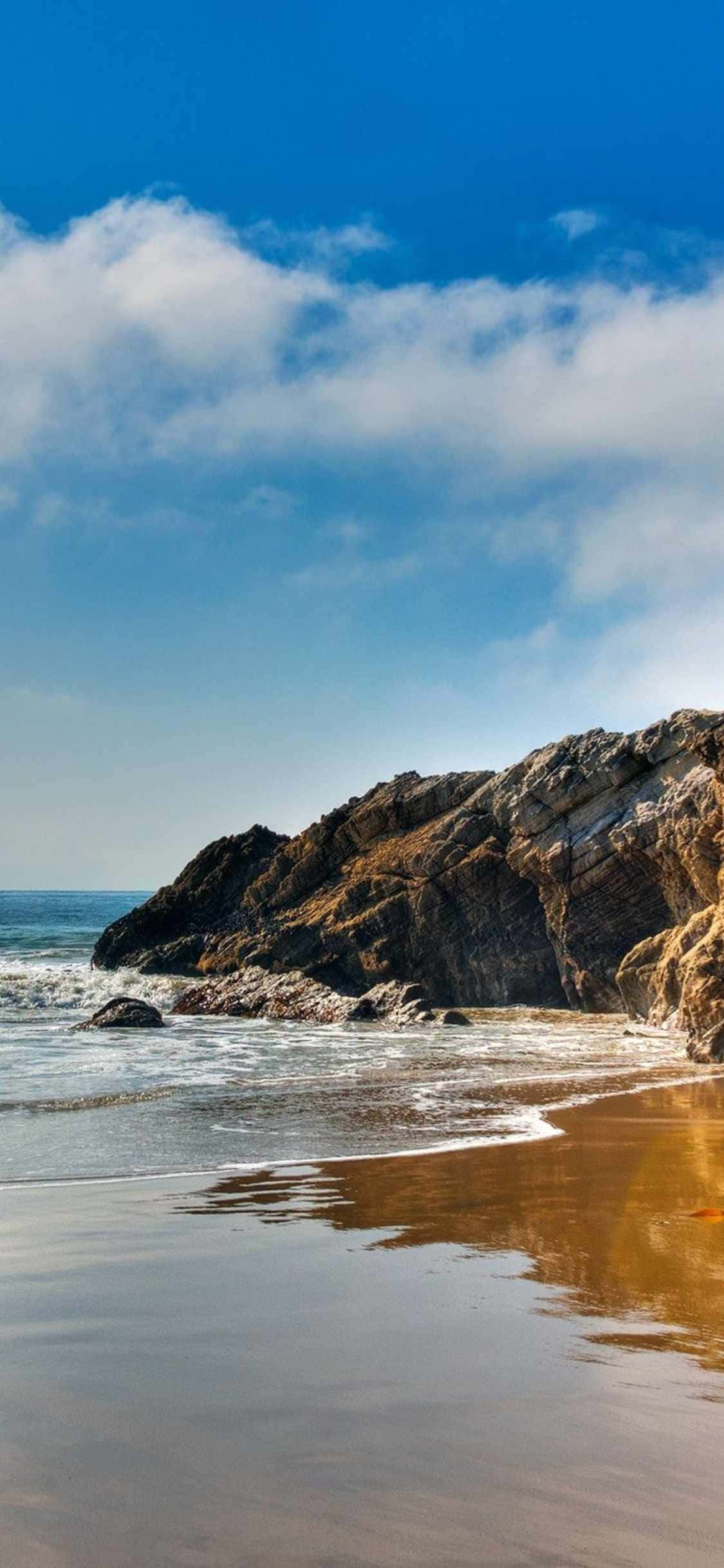 iPhone Xs Malibu 桌布 壁紙 — 陽光明媚的海灘，金色沙灘和拍打崎嶇懸崖的海浪，在明亮的藍天下有蓬鬆的白雲。