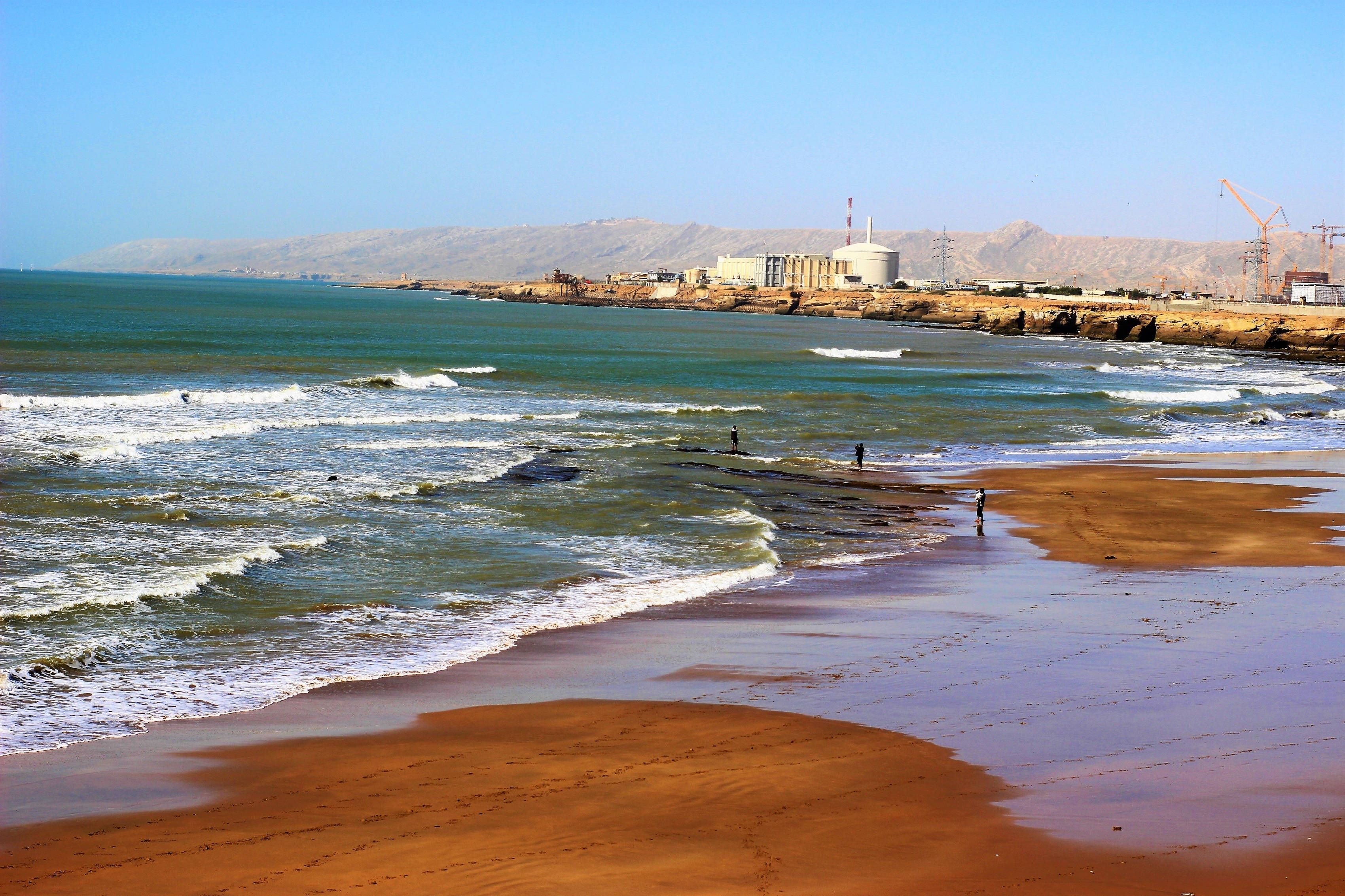 Karachi 桌布 壁紙 — 陽光明媚的海灘，金色沙灘和碧綠色的海浪在工業建築和遠處山脈附近拍打，天空清澈湛藍。