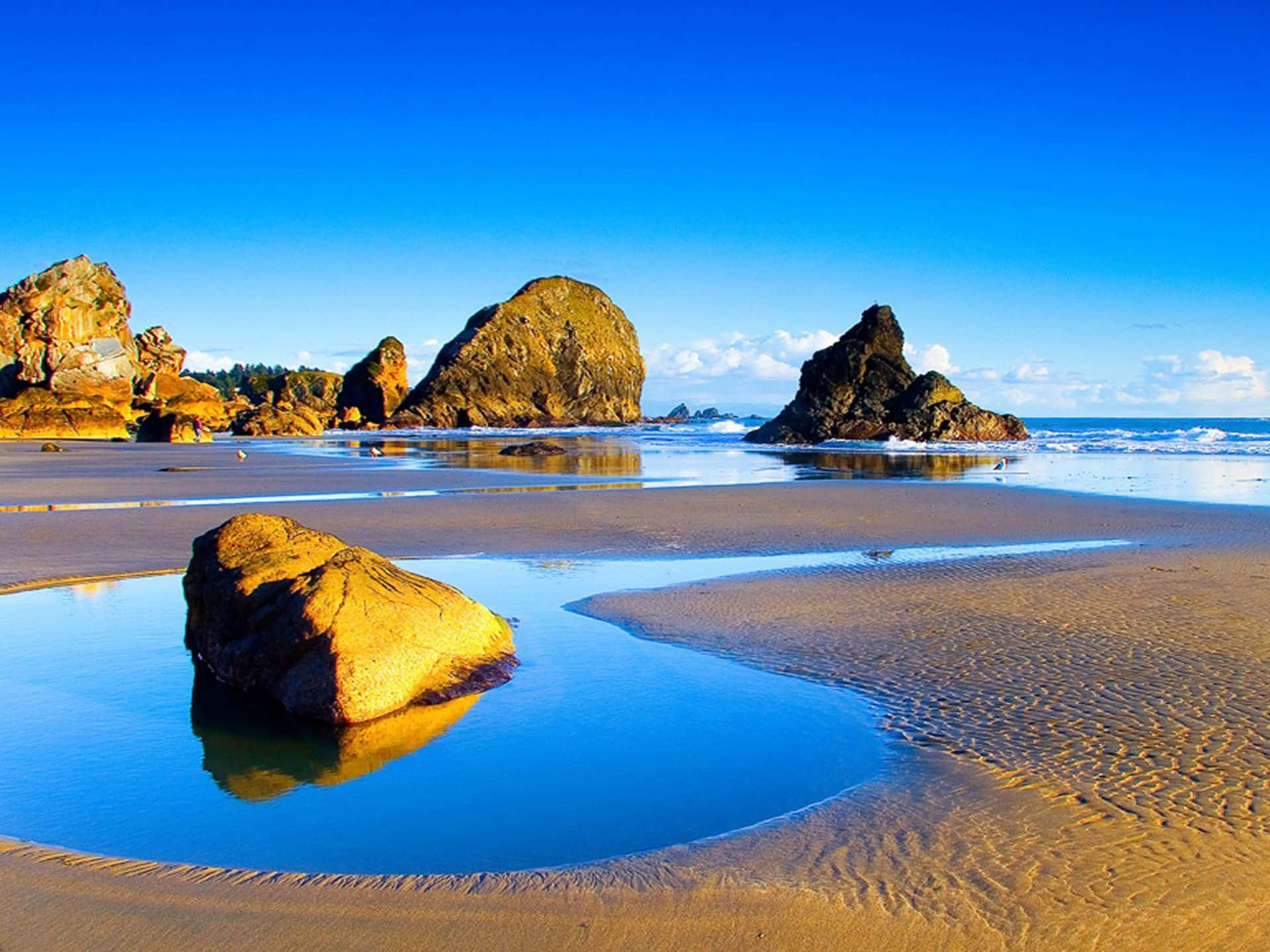 Beautiful Sea 桌布 壁紙 — 陽光明媚的海灘，金色沙紋和巨石在清澈藍天下的潮汐池中倒映。
