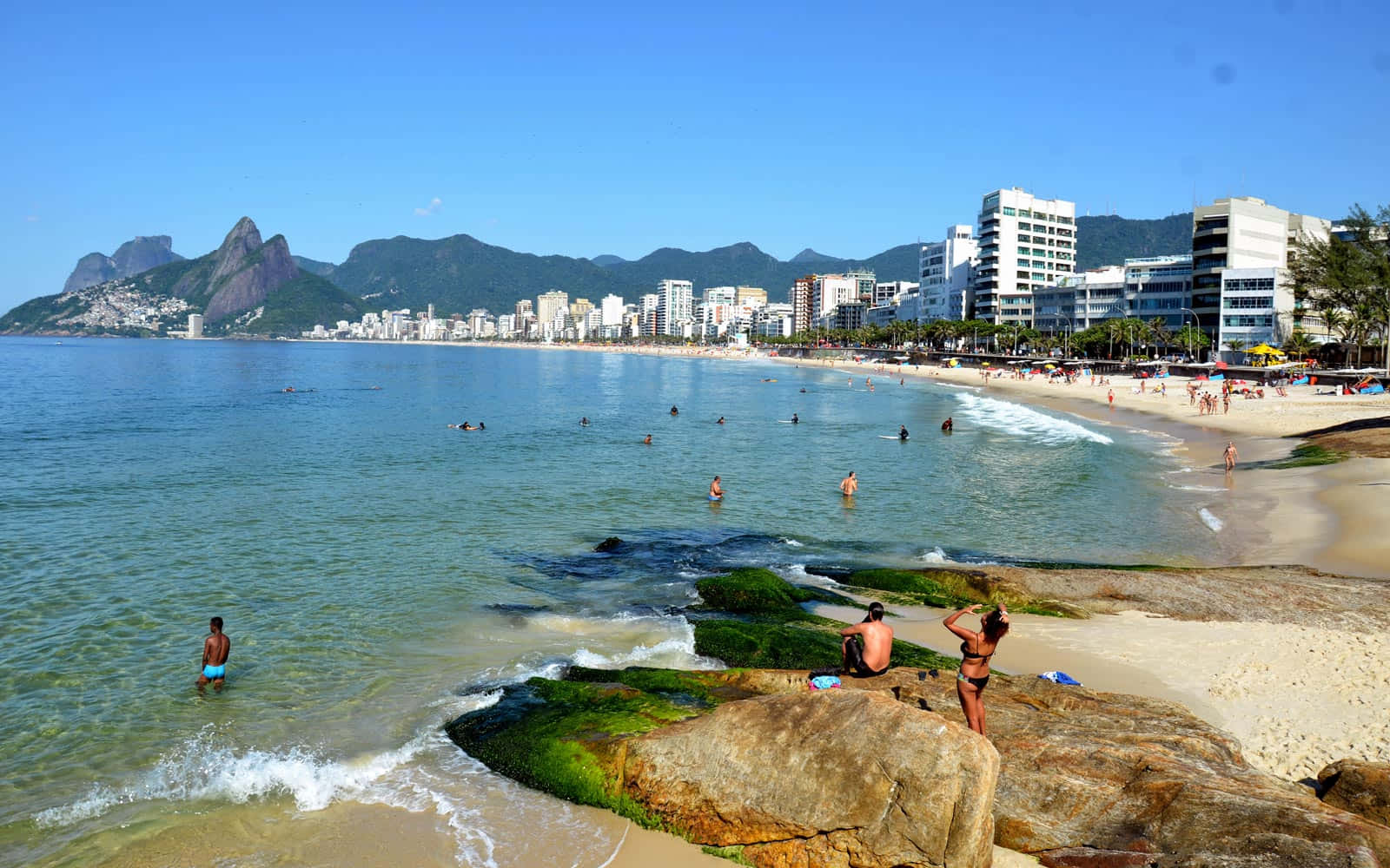 Brazilian Beach 桌布 壁紙 — 陽光明媚的海灘場景，人們在岩石海岸附近游泳和曬太陽，現代建築和標誌性的山脈在遠處，天空清澈湛藍。