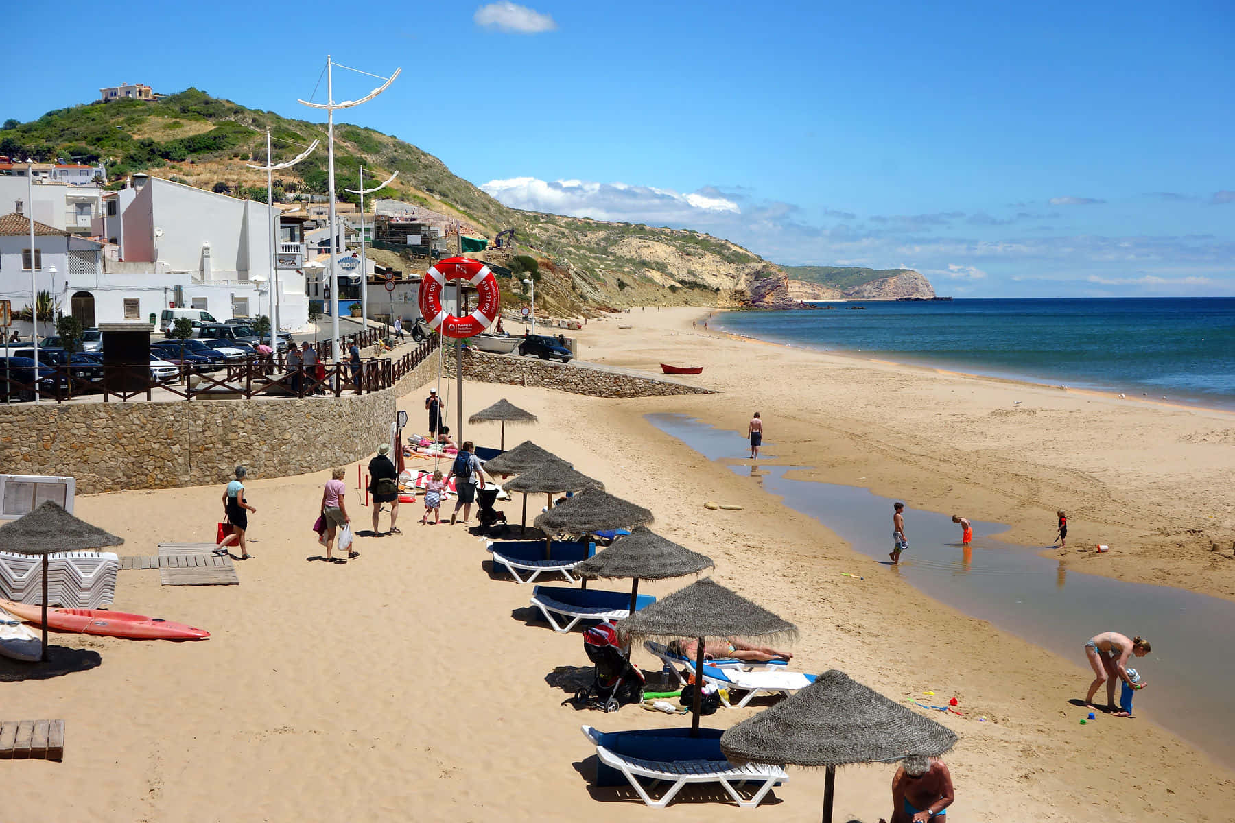Portuguese Beach 桌布 壁紙 — 陽光明媚的海灘場景，人們在草傘下放鬆，孩子們在水邊玩耍。