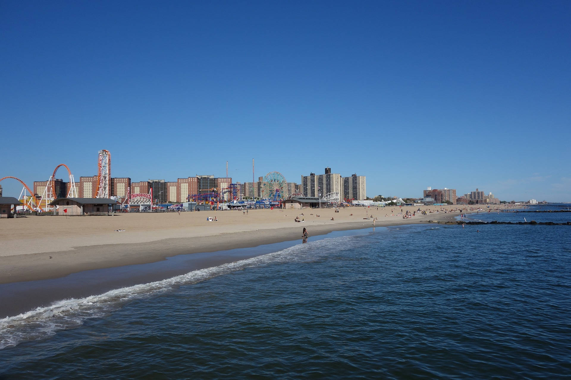 海灘 桌布 壁紙 — 陽光明媚的海灘場景，海浪翻滾，高樓後面有一個充滿活力的遊樂園，有雲霄飛車和摩天輪。