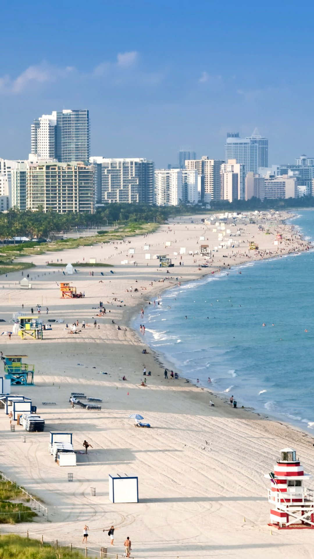 Miami iPhone 桌布 壁紙 — 陽光明媚的海灘場景，碧綠的海浪拍打著寬闊的沙灘，沙灘上排列著救生員塔，遠處的城市天際線在清澈的藍天下。