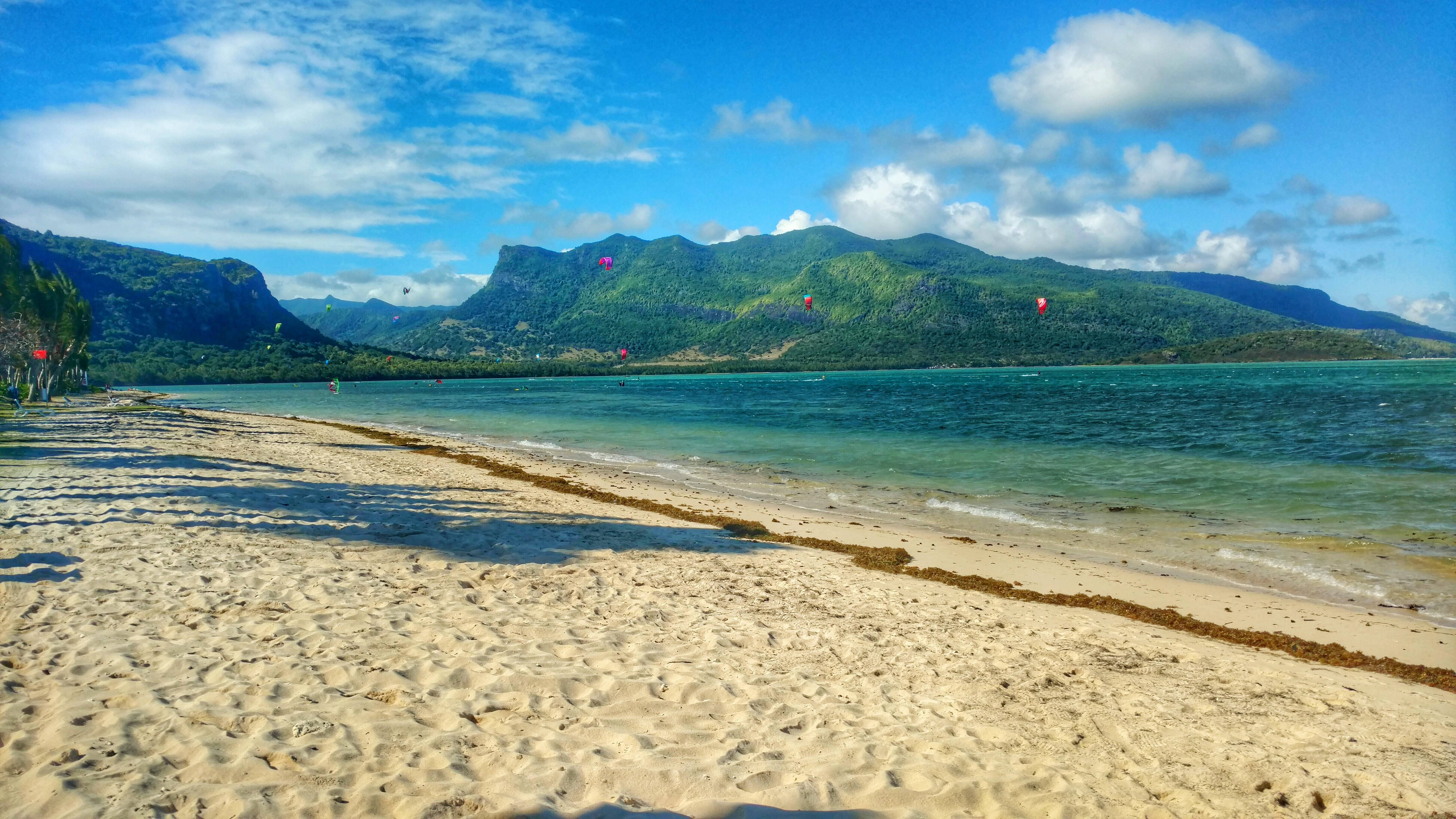Mauritius 桌布 壁紙 — 陽光明媚的熱帶海灘，金色沙灘和碧綠海水延伸至鬱鬱蔥蔥的綠色山脈，上方是明亮的藍天和蓬鬆的白雲。