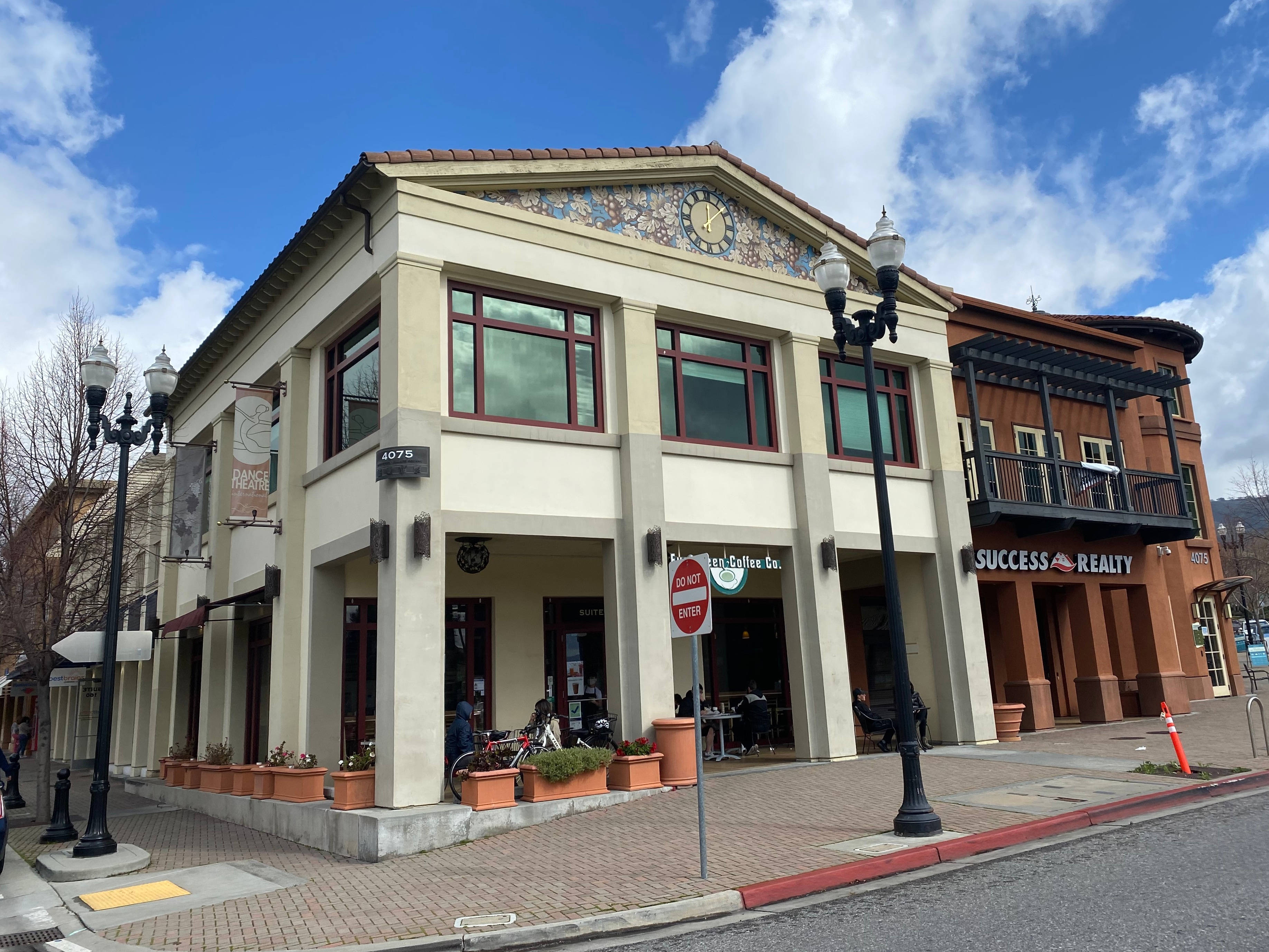 Village 桌布 壁紙 — 陽光明媚的街角，一棟米色建築內有咖啡店和房地產辦公室，上方是晴朗的藍天和蓬鬆的白雲。