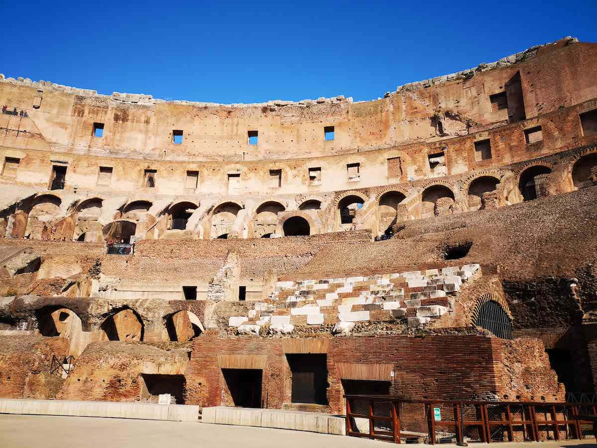 Roman Colosseum 桌布 壁紙 — 陽光普照的古羅馬競技場遺跡，風化的石拱門和上方清澈的藍天。