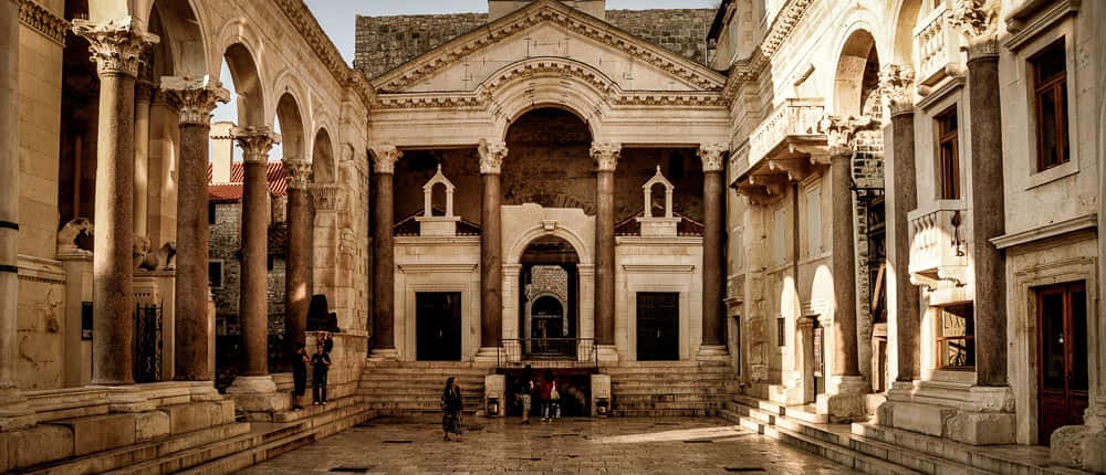 Croatia 桌布 壁紙 — 陽光普照的古老石頭庭院，宏偉的柱子和拱門在鵝卵石地面上投下長長的影子。