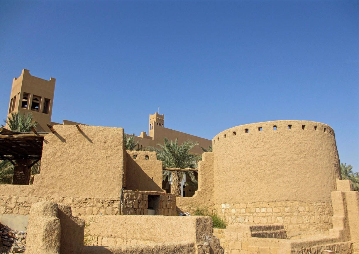 Riyadh 桌布 壁紙 — 陽光普照的土坯堡壘，圓形塔樓和棕櫚樹在清澈的藍天下。