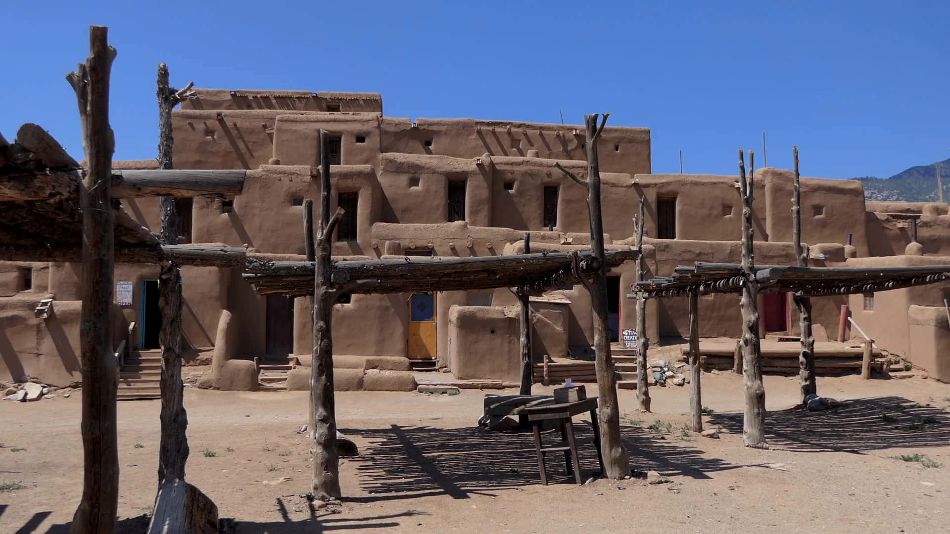 New Mexico 桌布 壁紙 — 陽光普照的土坯普韋布洛建築與木製涼棚，在晴朗藍天下於塵土飛揚的地面上投下長長的影子。