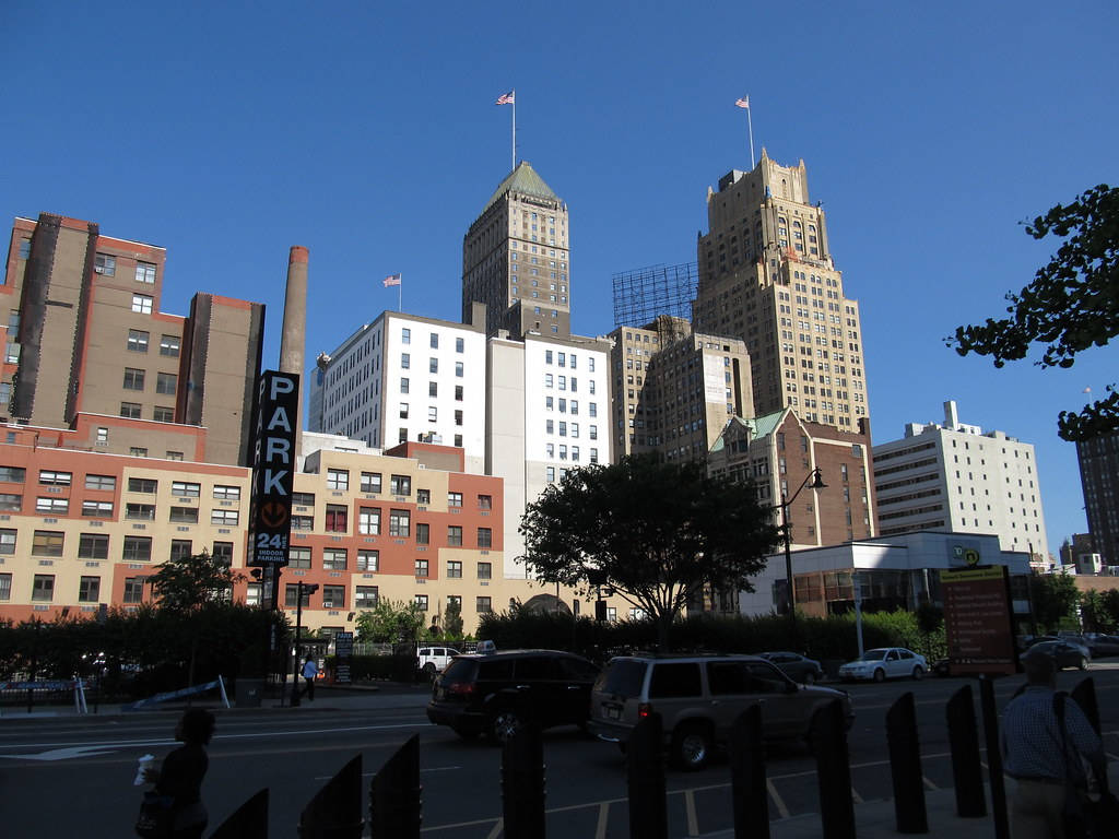 New Jersey 桌布 壁紙 — 陽光普照的城市天際線，歷史摩天大樓與現代建築在晴朗藍天下，下方有汽車和行人。