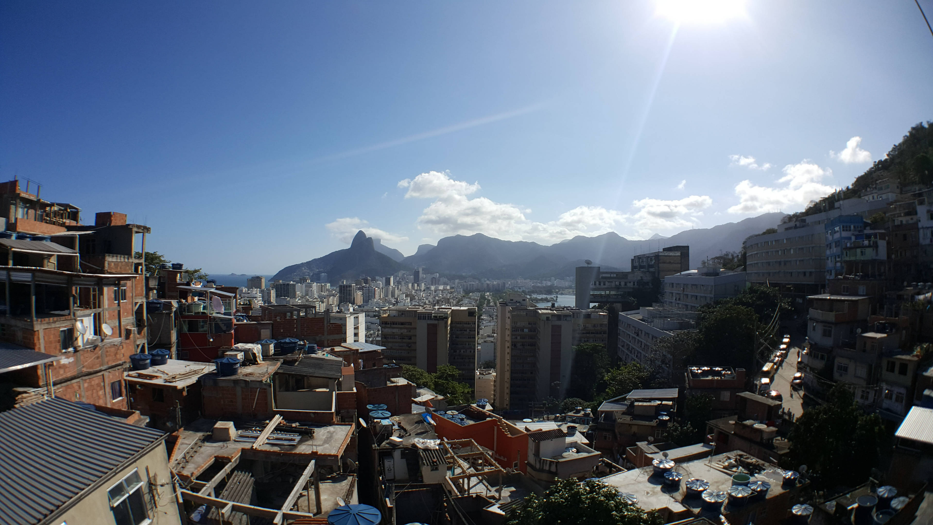 rio 桌布 壁紙 — 陽光普照的城市景觀，密集的紅磚建築沿著山丘向上延伸，俯瞰海灣和遠山，天空湛藍明亮。