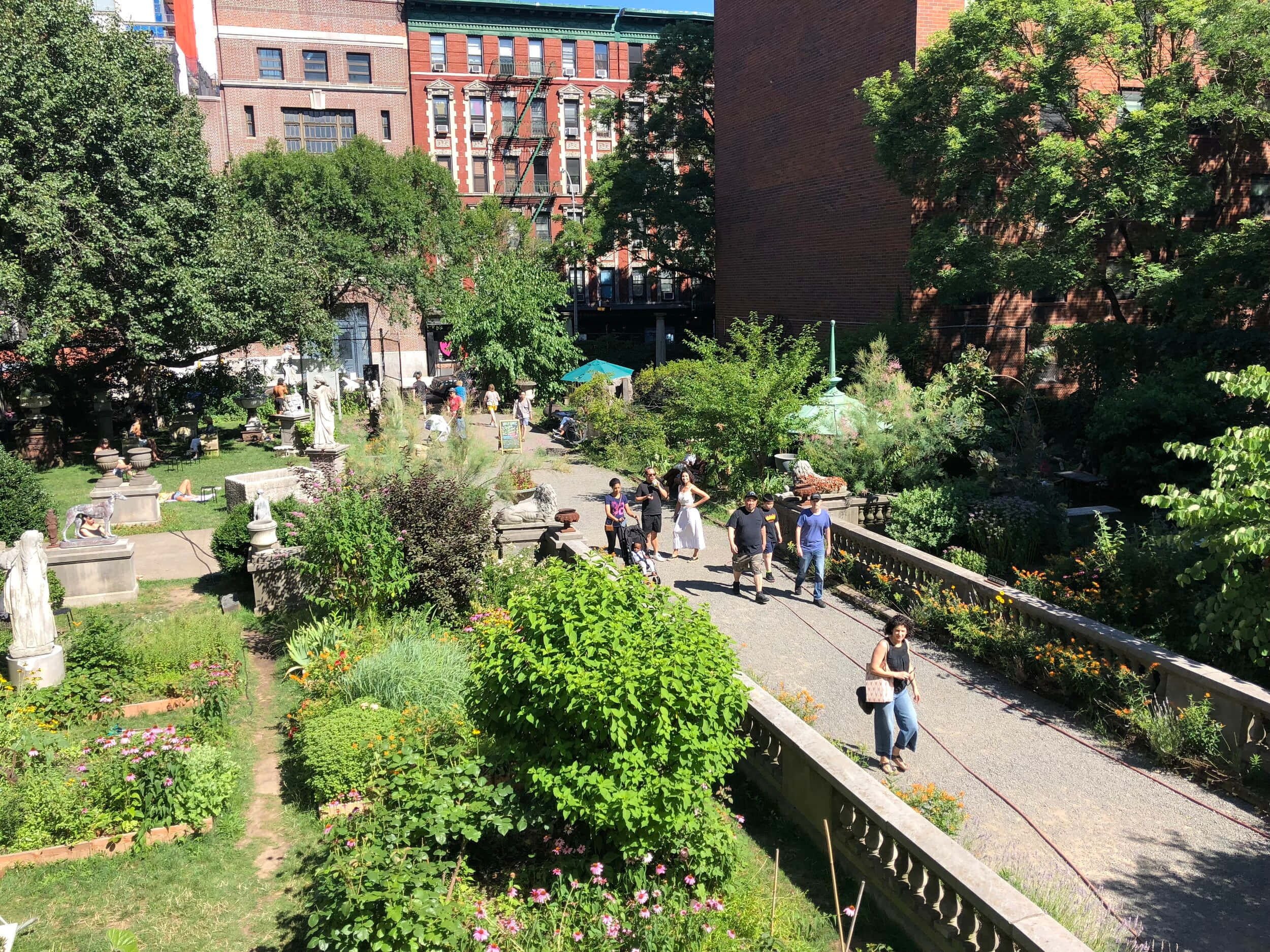 Street 桌布 壁紙 — 陽光普照的城市花園，人們在茂密綠植和古典雕像間漫步，四周環繞著磚砌建築。