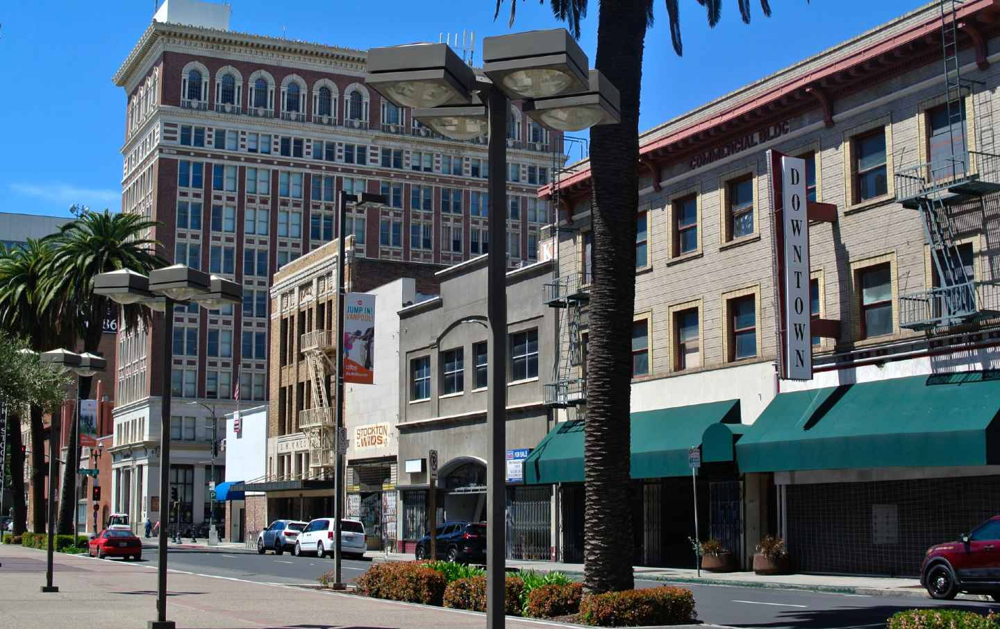 City Street 桌布 壁紙 — 陽光普照的市中心街道，兩旁有棕櫚樹、歷史建築和綠色遮陽篷，天空清澈湛藍。