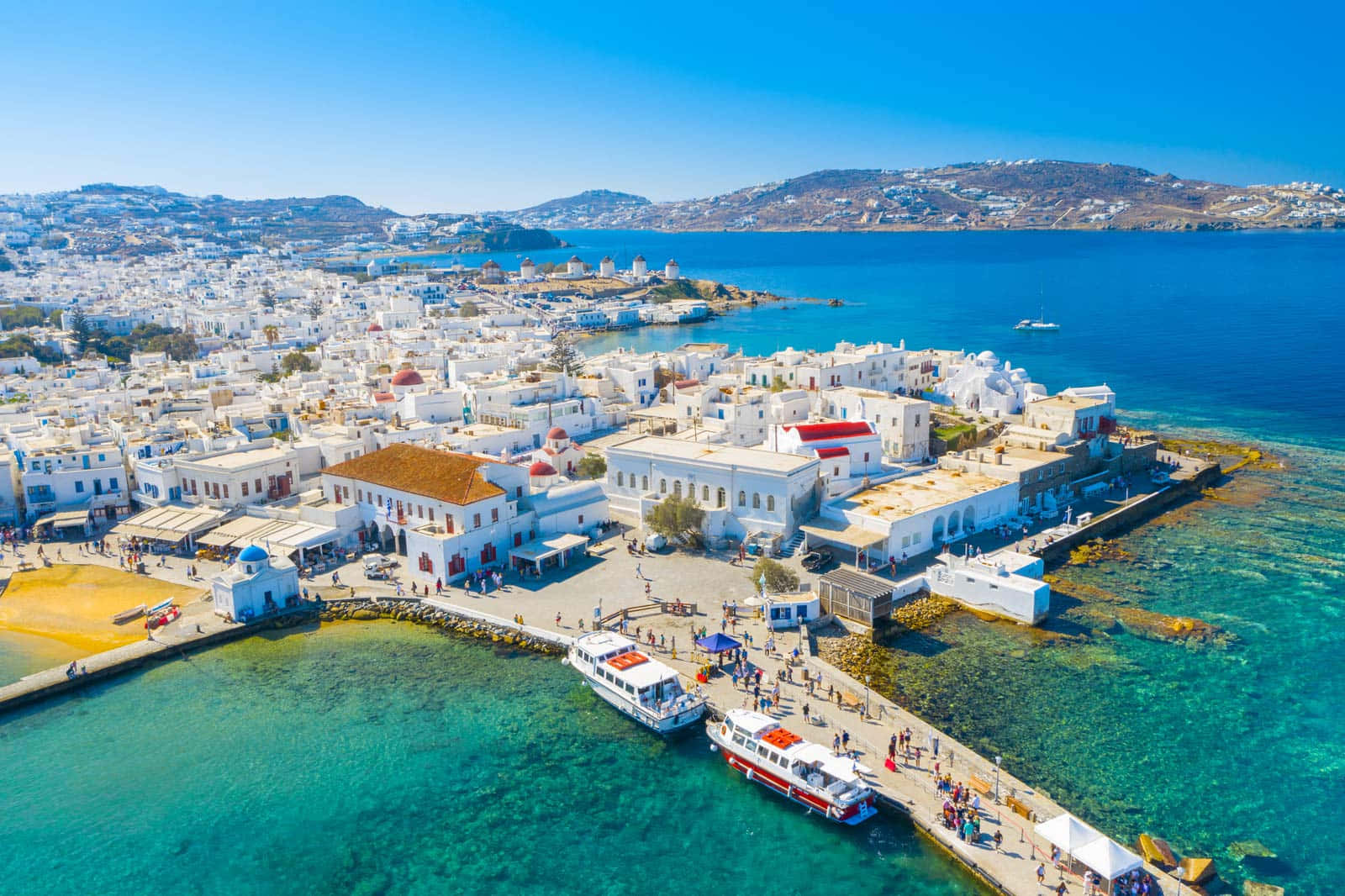 Europe 桌布 壁紙 — 陽光普照的希臘島嶼小鎮鳥瞰圖，白色建築環抱著碧綠海水，船隻停泊在繁忙的碼頭。