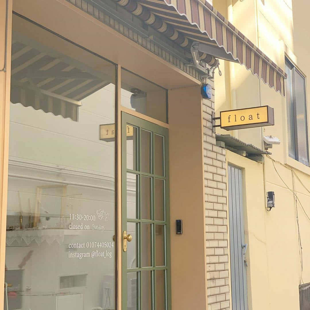 美學米色 桌布 壁紙 — 陽光普照的店鋪門面，帶有條紋遮陽篷和玻璃門，反射街道，黃色招牌上寫著 float。