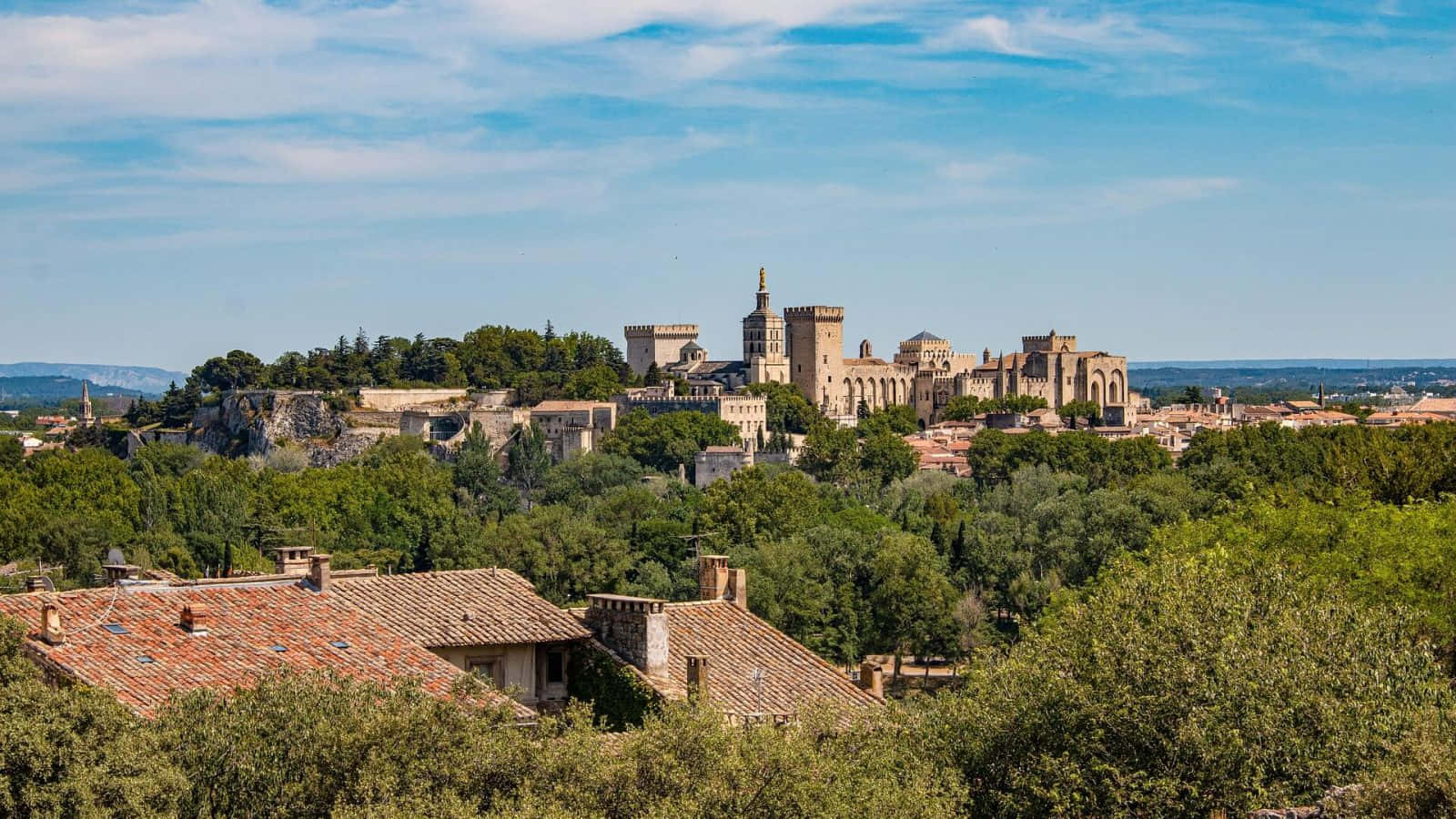 trees 桌布 壁紙 — 陽光普照的景色，一座歷史悠久的石城堡建築群坐落在山頂上，俯瞰下方綠意盎然的山谷和陶土屋頂。