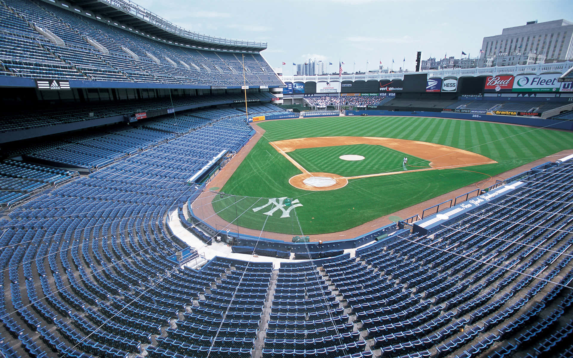 Baseball Field 桌布 壁紙 — 陽光普照的棒球場鳥瞰圖，空藍色座位環繞著一個帶有大型洋基隊標誌的充滿活力的綠色場地。