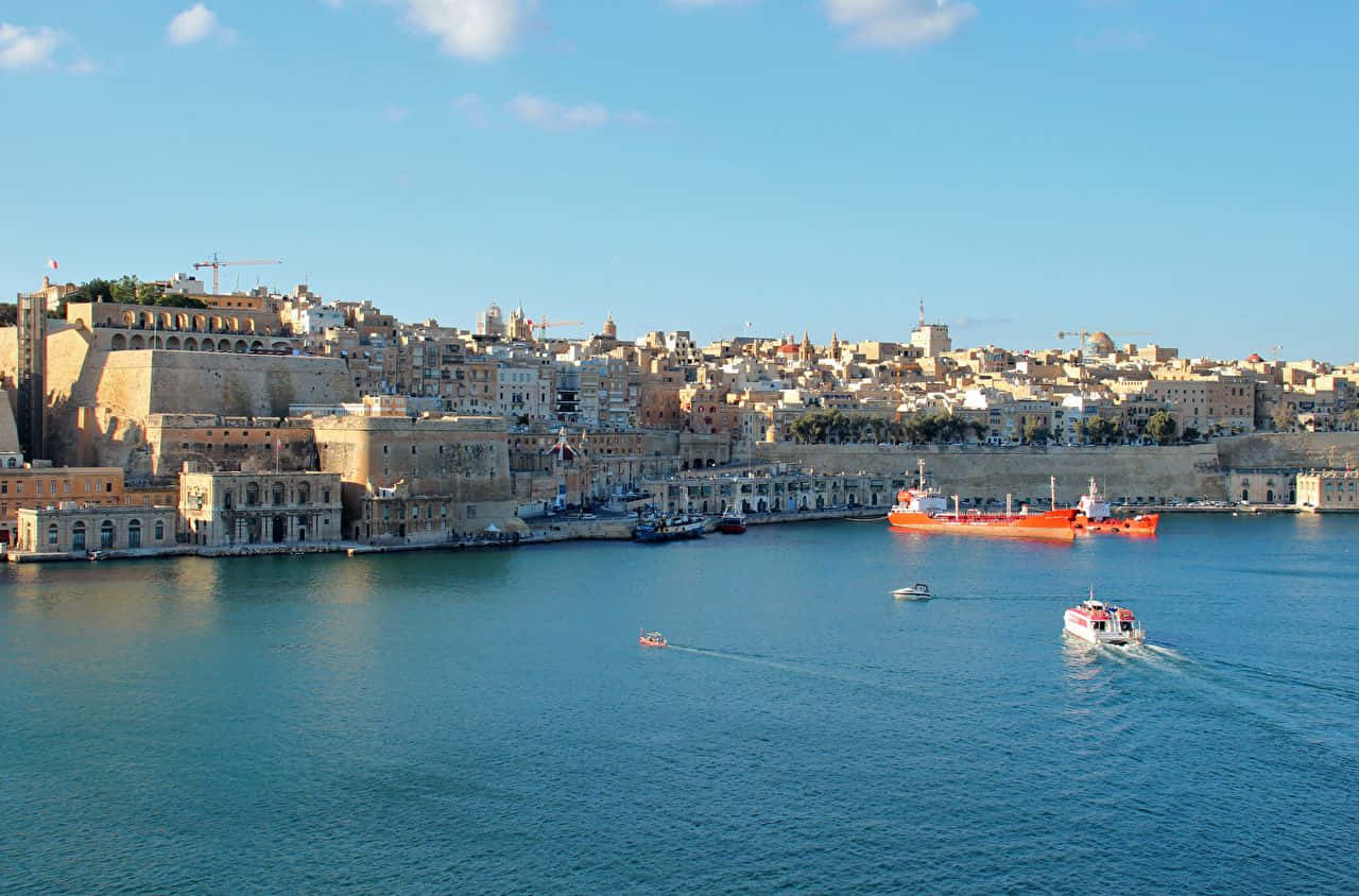 Malta 桌布 壁紙 — 陽光普照的歷史港口城市，石頭建築群聚在懸崖上，俯瞰平靜的藍色海水，點綴著船隻。