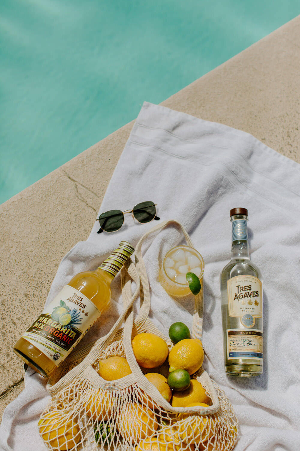 margaritas 桌布 壁紙 — 陽光普照的池畔場景，白色毛巾上放著一瓶龍舌蘭瑪格麗特混合飲料、檸檬、萊姆、太陽眼鏡和一杯雞尾酒。