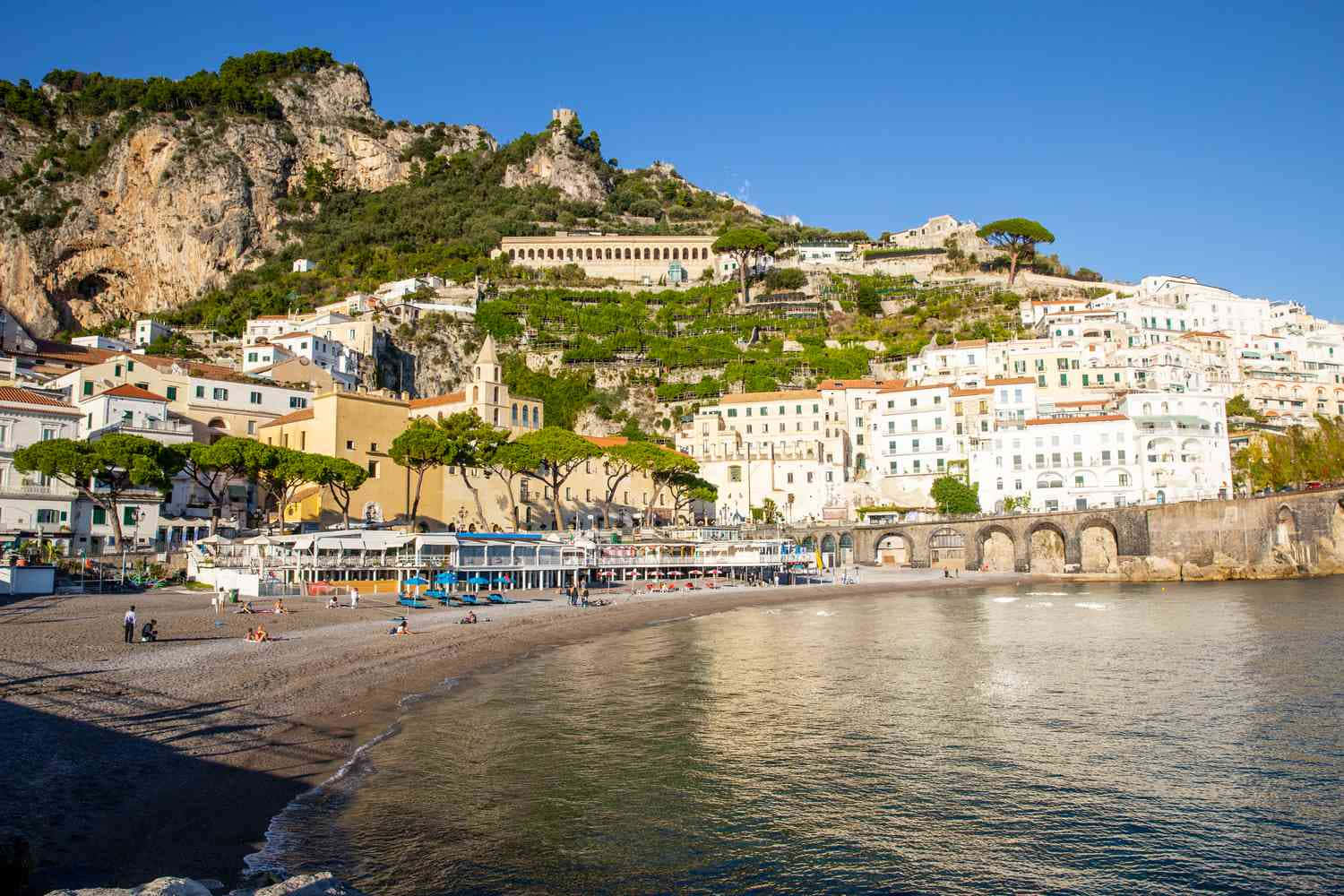 Italian Beach 桌布 壁紙 — 陽光普照的海岸小鎮，白色建築物沿懸崖層疊而下，通往卵石海灘和風平浪靜的大海，天空清澈湛藍。