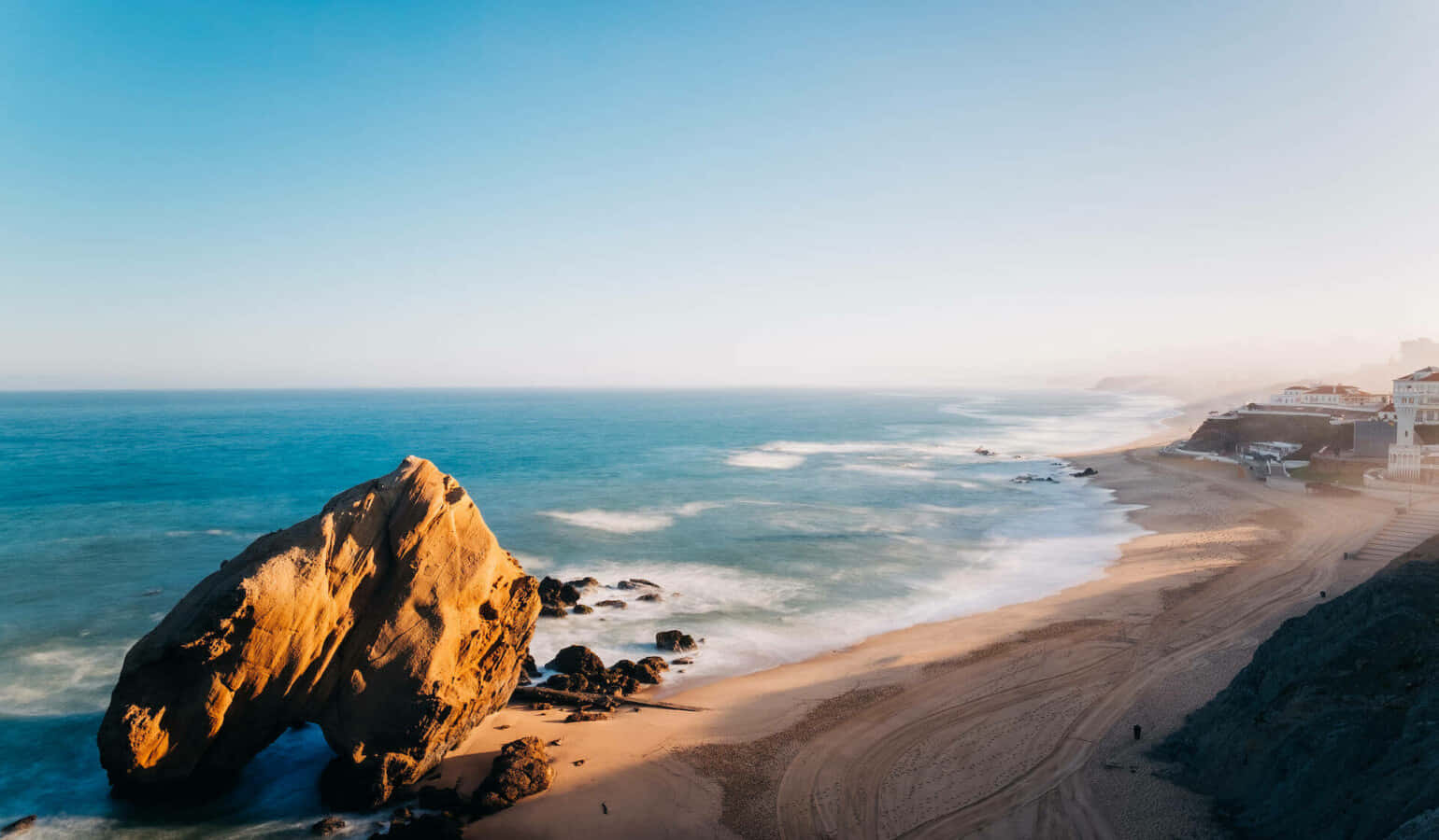 Portuguese Beach 桌布 壁紙 — 陽光普照的海岸景色，沙灘旁碧綠海水中矗立著巨大的金色岩石。
