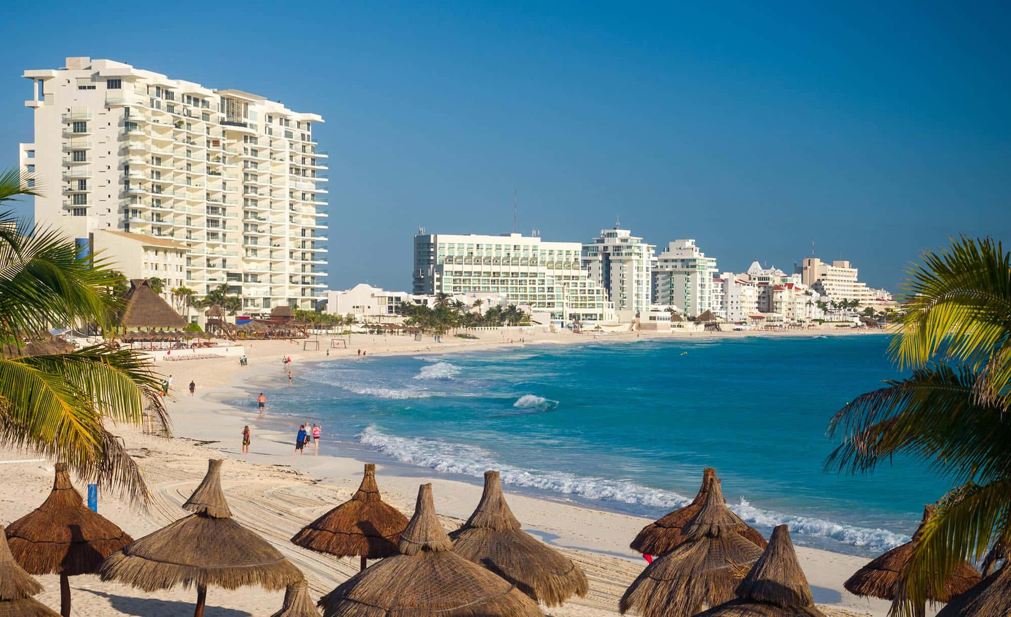 Mexican Beach 桌布 壁紙 — 陽光普照的海濱，碧綠的海浪拍打著白色沙灘，沙灘上排列著茅草傘，高聳的白色度假酒店在清澈的藍天下。