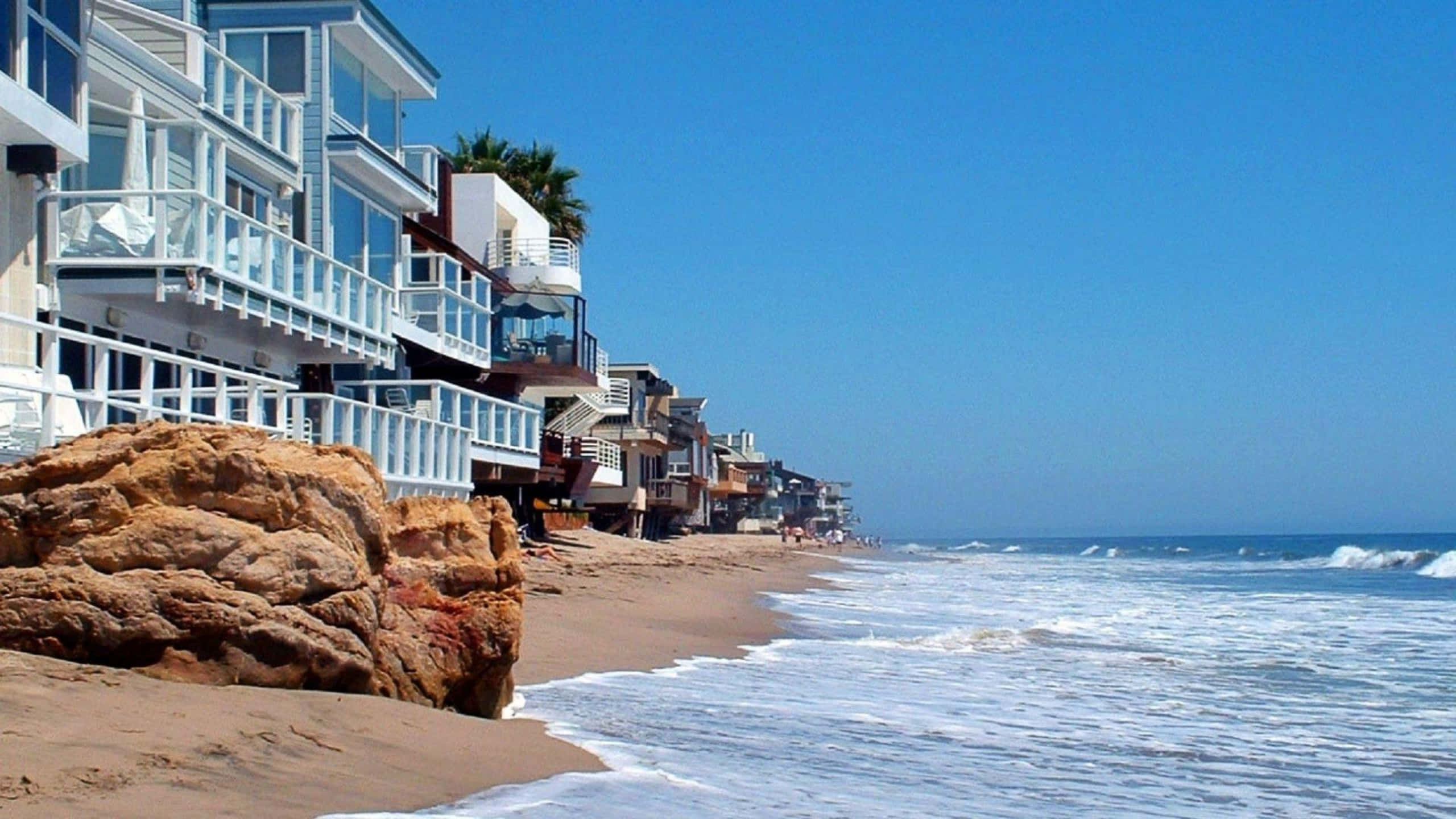 Beach House 桌布 壁紙 — 陽光普照的海濱房屋，白色陽台沿著沙灘排列，海浪在清澈的藍天下拍打岩石。
