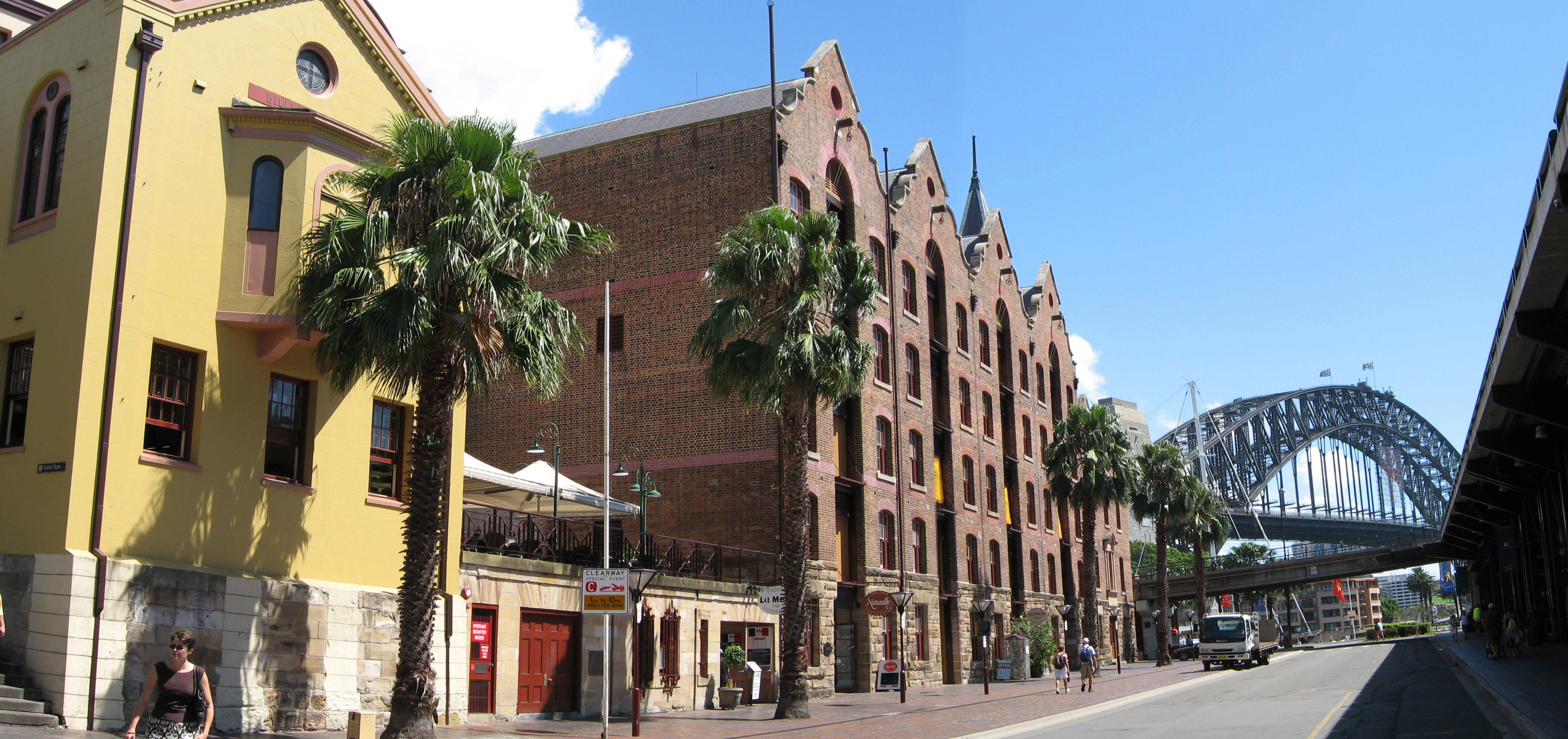 Sydney 桌布 壁紙 — 陽光普照的海濱街道，兩旁種有棕櫚樹和歷史悠久的磚砌建築，通往標誌性的雪梨港灣大橋。