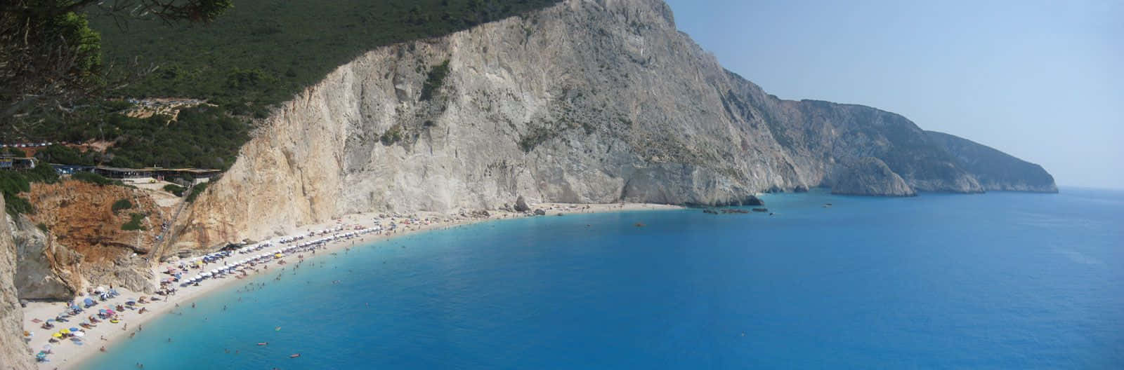 Greek Island 桌布 壁紙 — 陽光普照的海灘全景，碧綠的海水 nestled 在高聳的岩石峭壁和茂密的綠植之間。