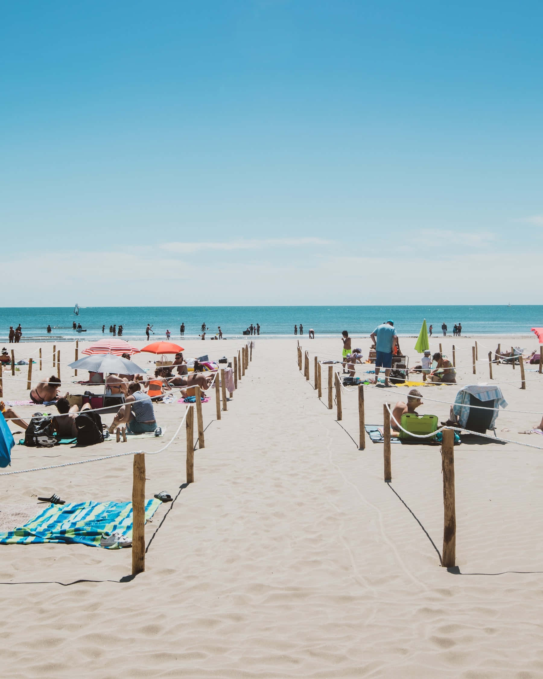 French Beach 桌布 壁紙 — 陽光普照的海灘場景，人們在色彩繽紛的遮陽傘下放鬆，木樁劃分出通往海洋的沙路。