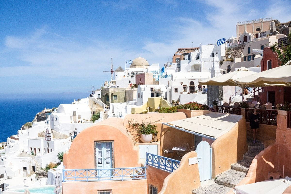 Santorini 桌布 壁紙 — 陽光普照的白色建築物沿著懸崖層層疊疊，俯瞰著晴朗天空下深藍色的愛琴海。