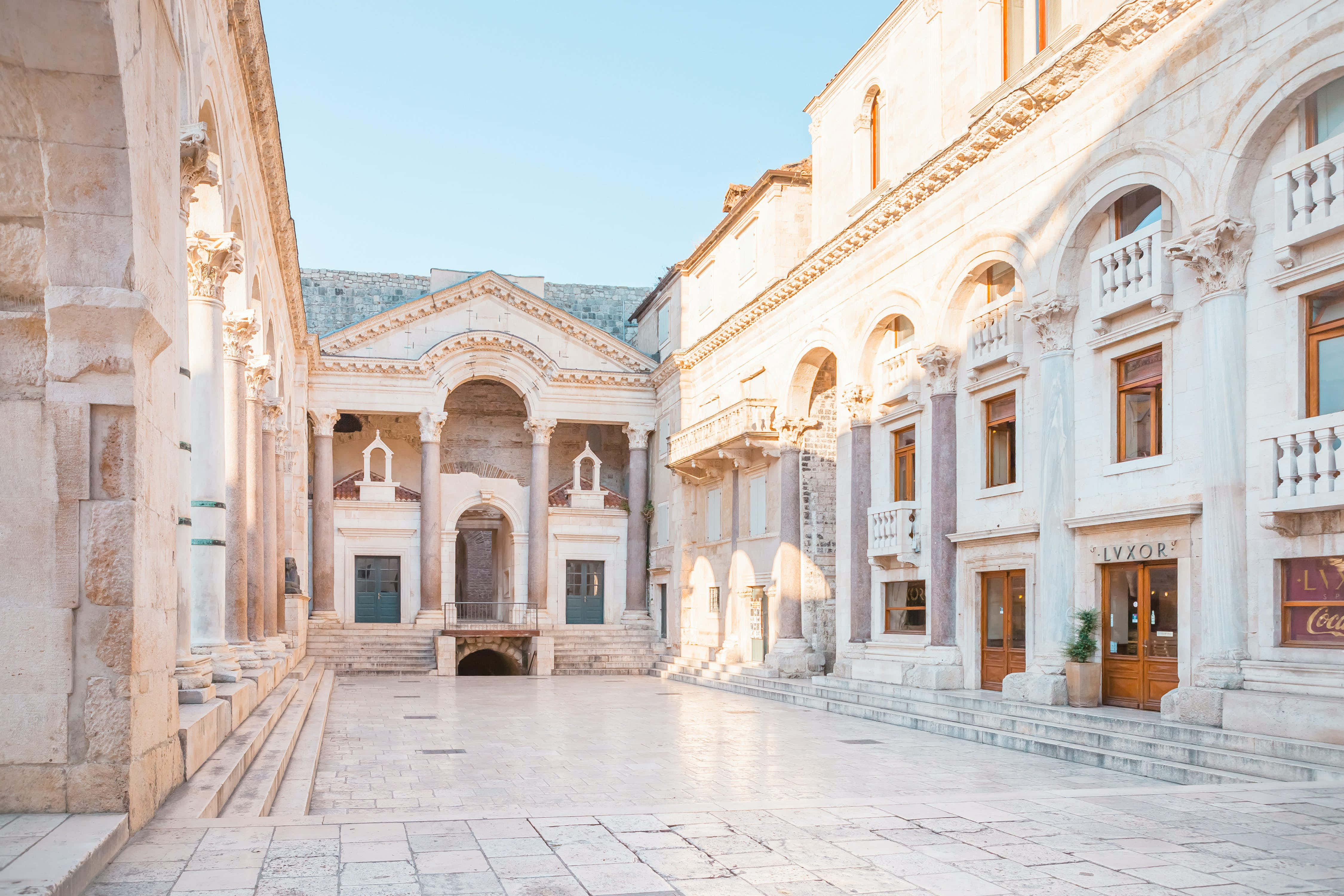 Croatia 桌布 壁紙 — 陽光普照的石頭庭院，以古典建築、拱形柱廊和中央神廟式結構為框架。