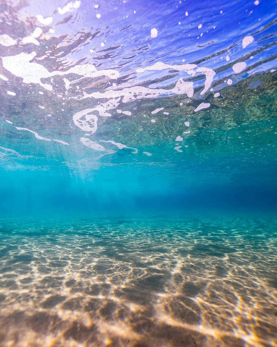 海灘 桌布 壁紙 — 陽光普照的碧綠海洋水下景觀，漣漪般的光影在沙質海底和上方泡沫表面舞動。