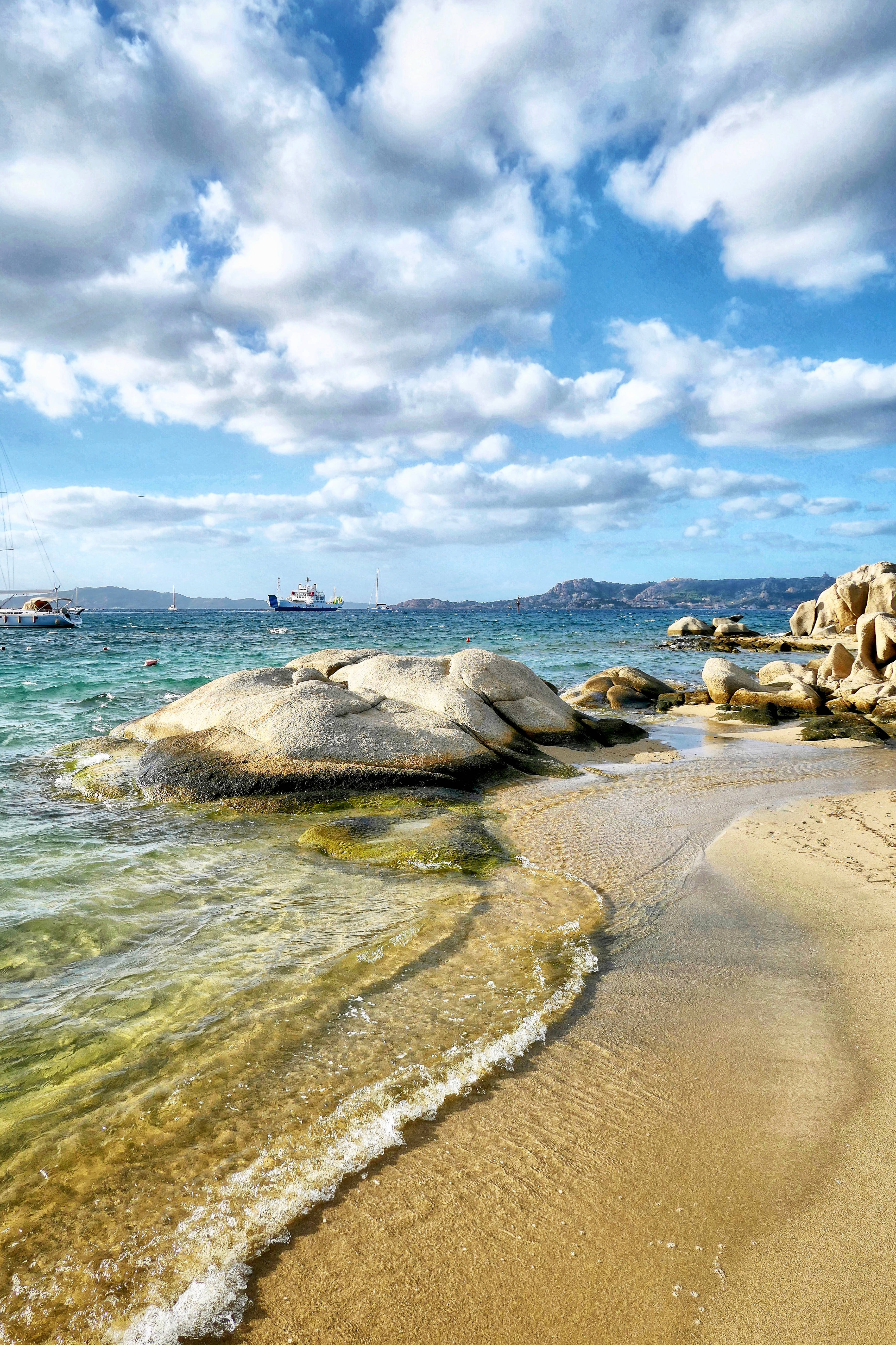Palau 桌布 壁紙 — 陽光普照的碧綠海浪拍打著金色沙灘，旁邊是風化的岩石，上方是點綴著蓬鬆白雲的明亮藍天。