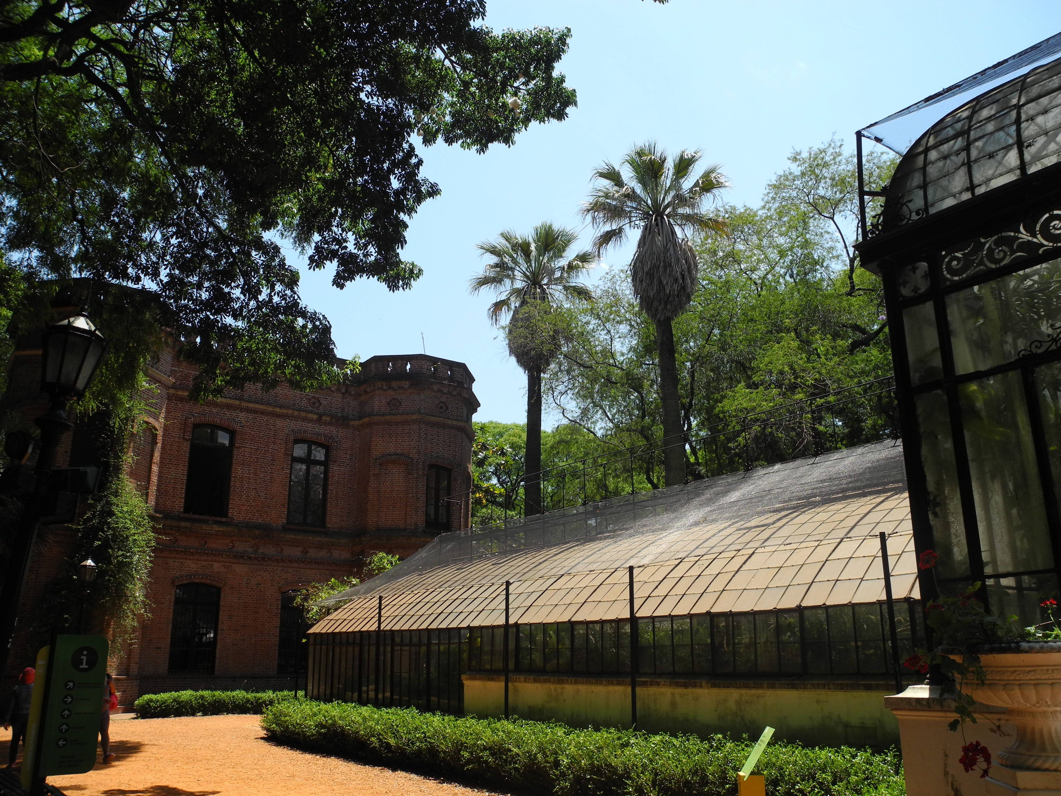 Aires 桌布 壁紙 — 陽光普照的花園場景，設有紅磚建築、高大的棕櫚樹和玻璃溫室，在晴朗的藍天下。