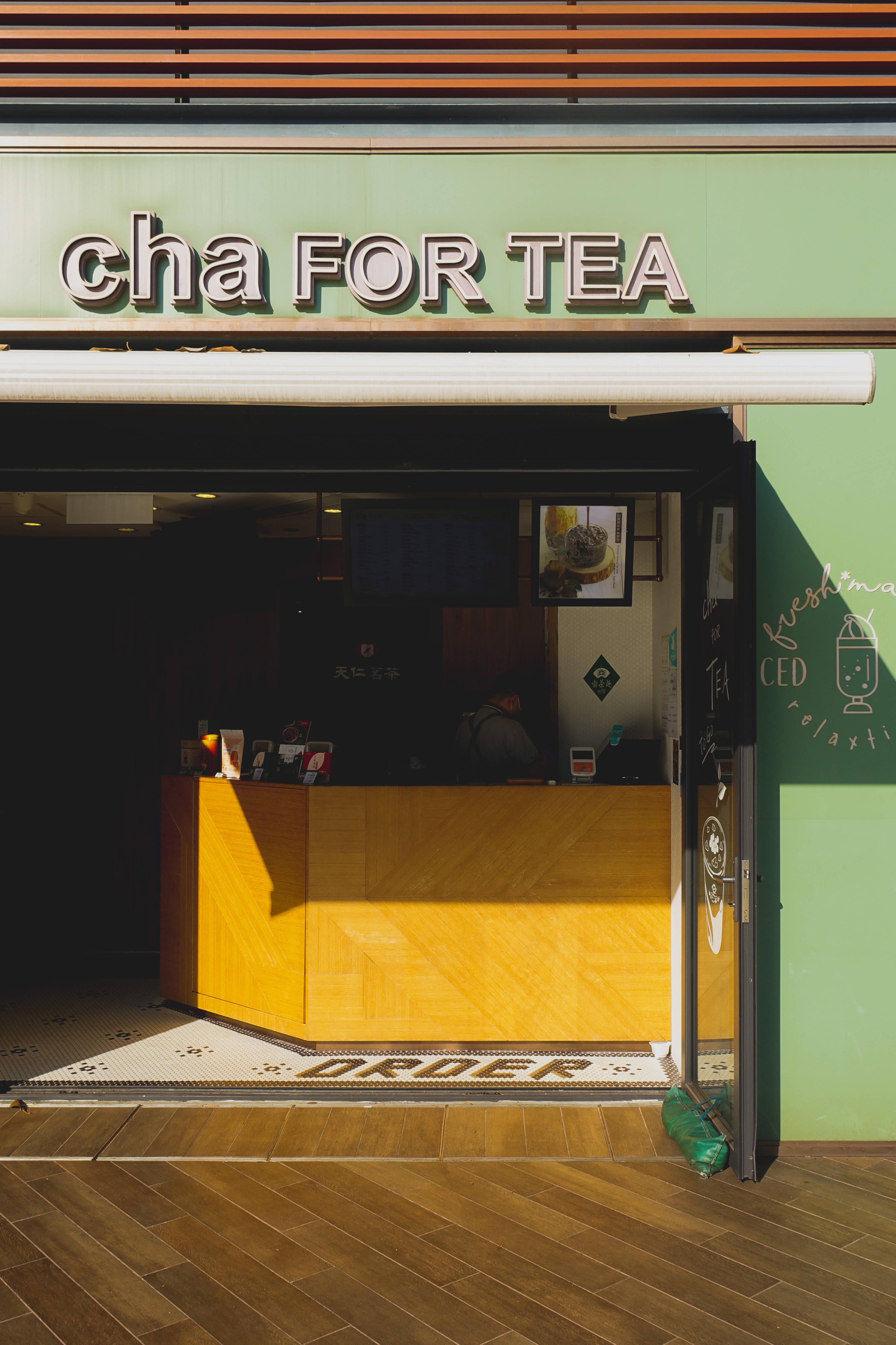 綠色美學 桌布 壁紙 — 陽光普照的茶店入口，綠色招牌上寫著 cha FOR TEA，木製櫃檯投下溫暖的陰影。