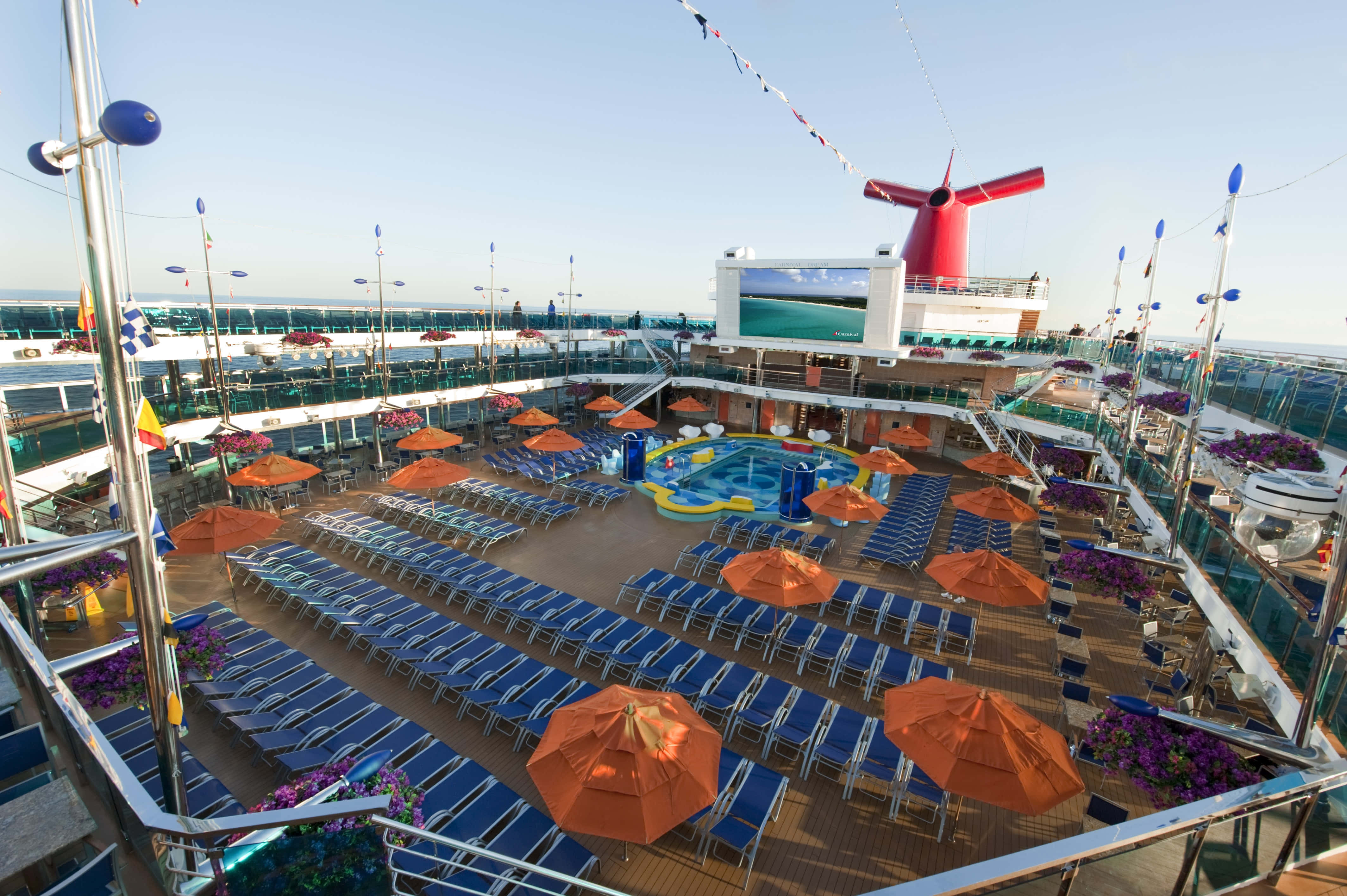 Carnival Dream 桌布 壁紙 — 陽光普照的郵輪甲板，排列著藍色躺椅、橙色遮陽傘和色彩繽紛的泳池，天空晴朗。