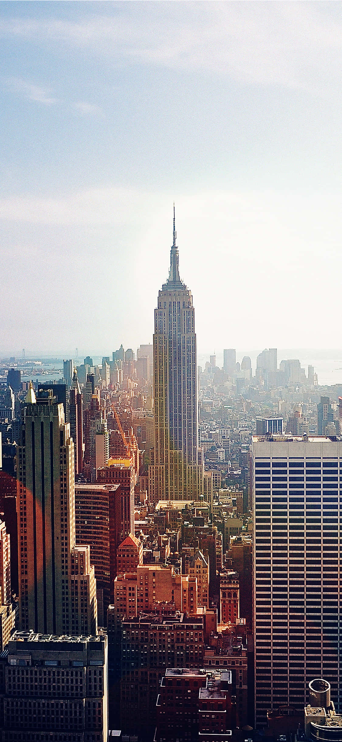 New York 桌布 壁紙 — 陽光沐浴著標誌性的帝國大廈，它在晴朗的藍天下聳立在密集的紐約市天際線之上。
