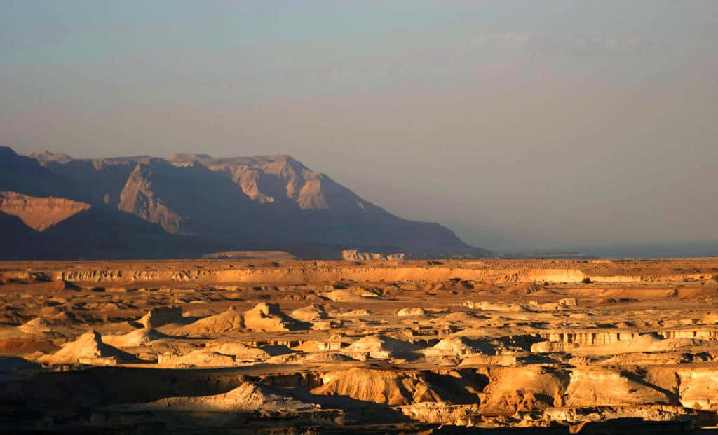Israel 桌布 壁紙 — 陽光灑落在一個崎嶇的沙漠峽谷，峽谷中有層狀岩層和遠山，天空朦朧。