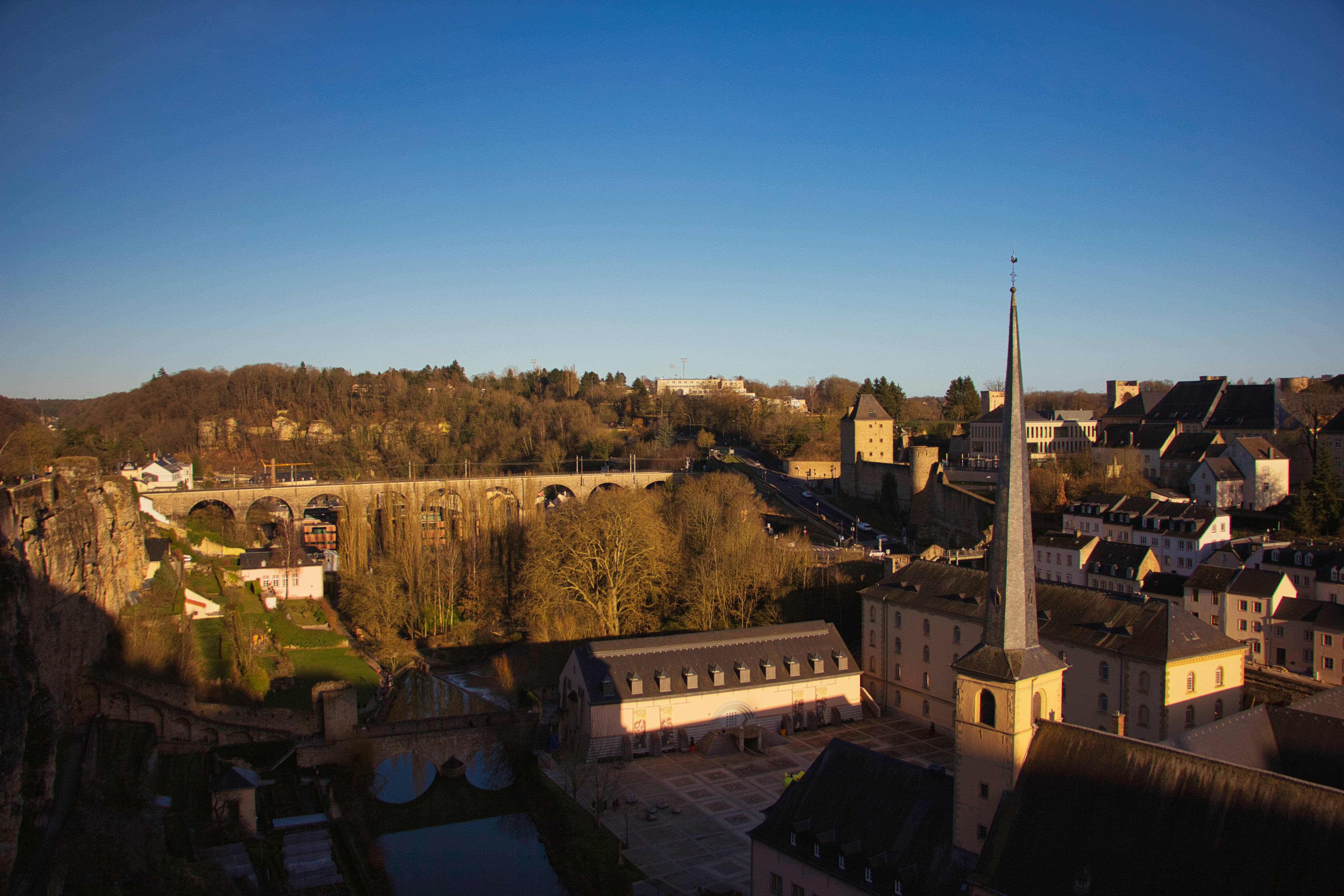 Luxembourg 桌布 壁紙 — 陽光灑落在一個歷史悠久的歐洲小鎮，鎮上有石橋和教堂尖塔，背景是清澈的藍天。