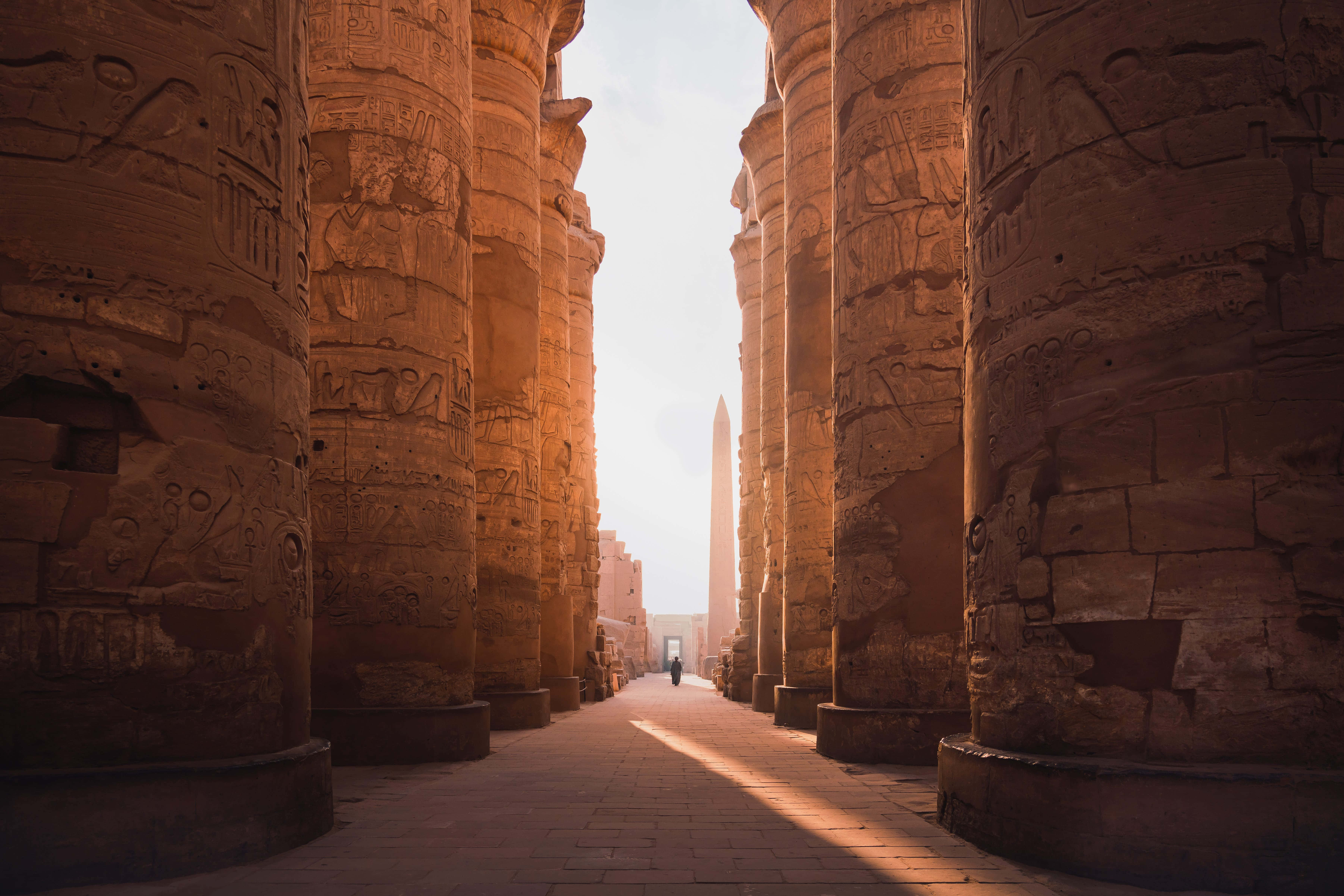Ancient Egypt 桌布 壁紙 — 陽光灑落在刻有象形文字的宏偉古埃及柱子上，一個孤獨的身影正走向遠處的方尖碑。