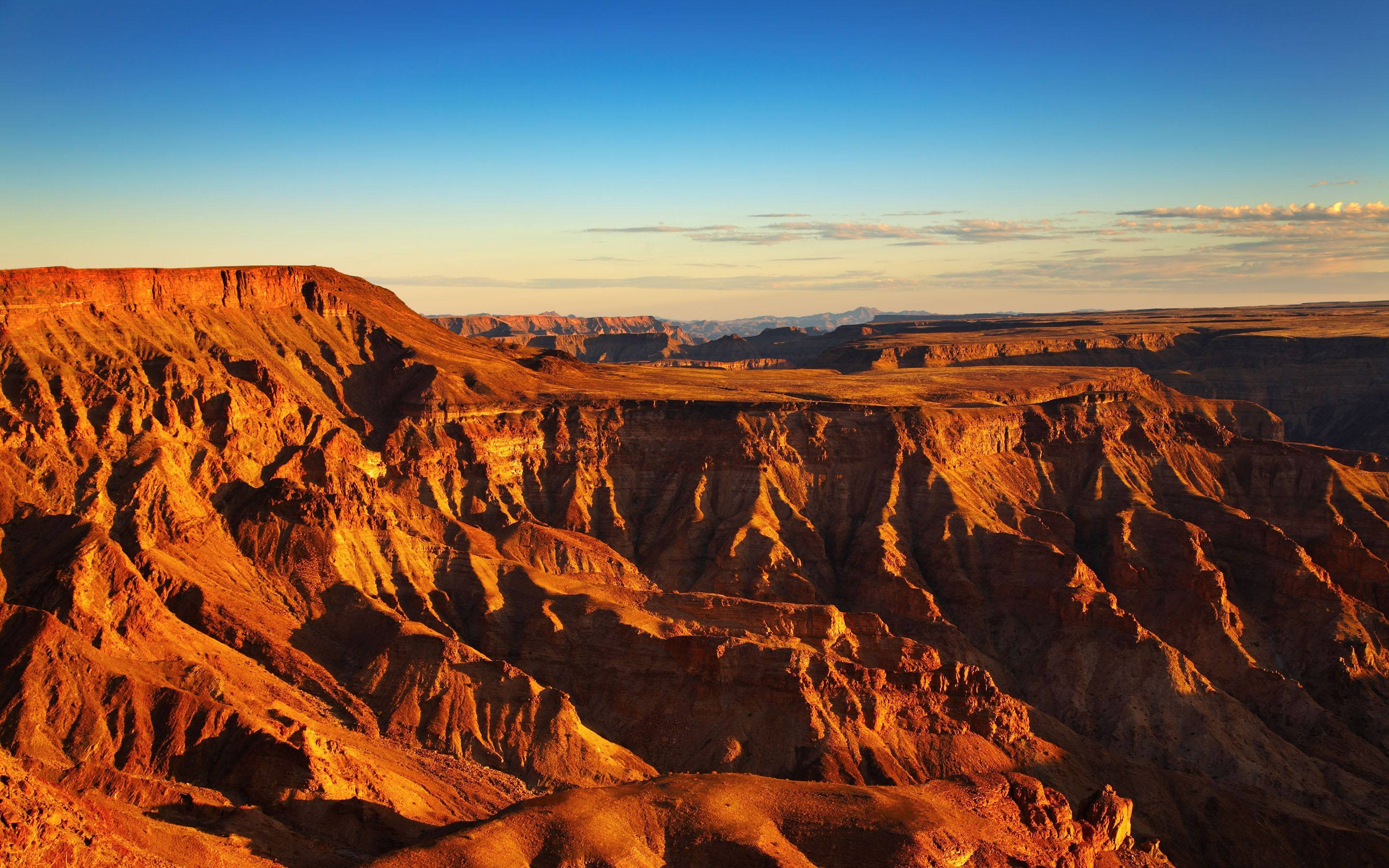 Namibia 桌布 壁紙 — 陽光灑落在廣闊沙漠景觀中崎嶇的紅色懸崖和深邃峽谷上，天空清澈湛藍。
