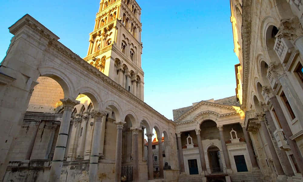 Croatia 桌布 壁紙 — 陽光灑落在歷史悠久的歐洲庭院中，古老的石拱門和高聳的鐘樓映襯著清澈的藍天。