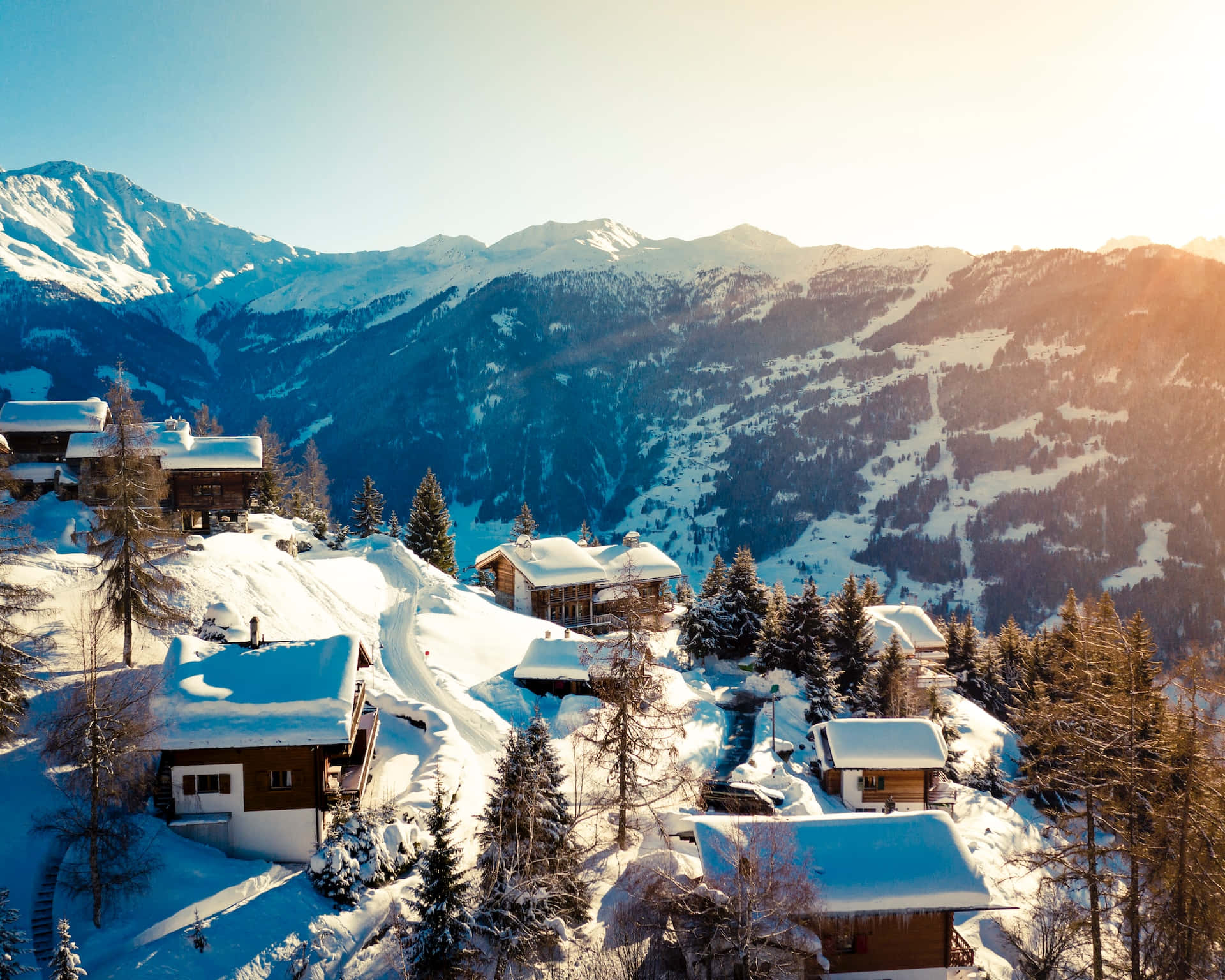 Ski Resort 桌布 壁紙 — 陽光灑落在被雪覆蓋的阿爾卑斯山小木屋上，這些小木屋隱藏在常青樹之間，背景是雄偉的雪山。