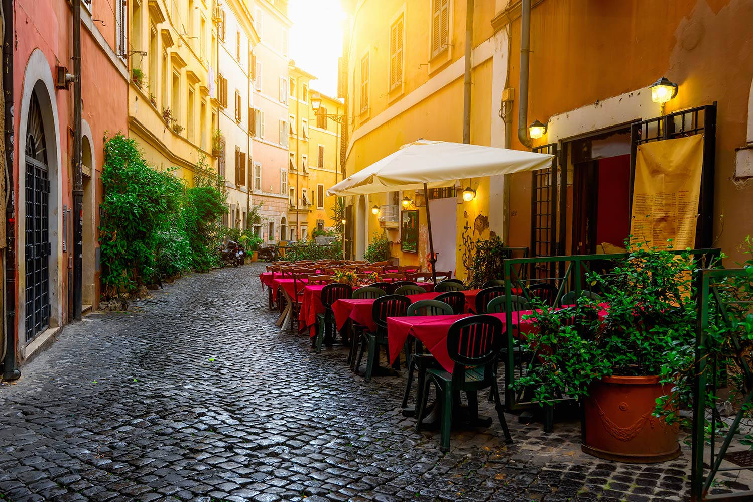 Rome 桌布 壁紙 — 陽光照亮了一條狹窄的鵝卵石小巷，兩旁是色彩繽紛的建築物和鋪著紅布的戶外咖啡桌。
